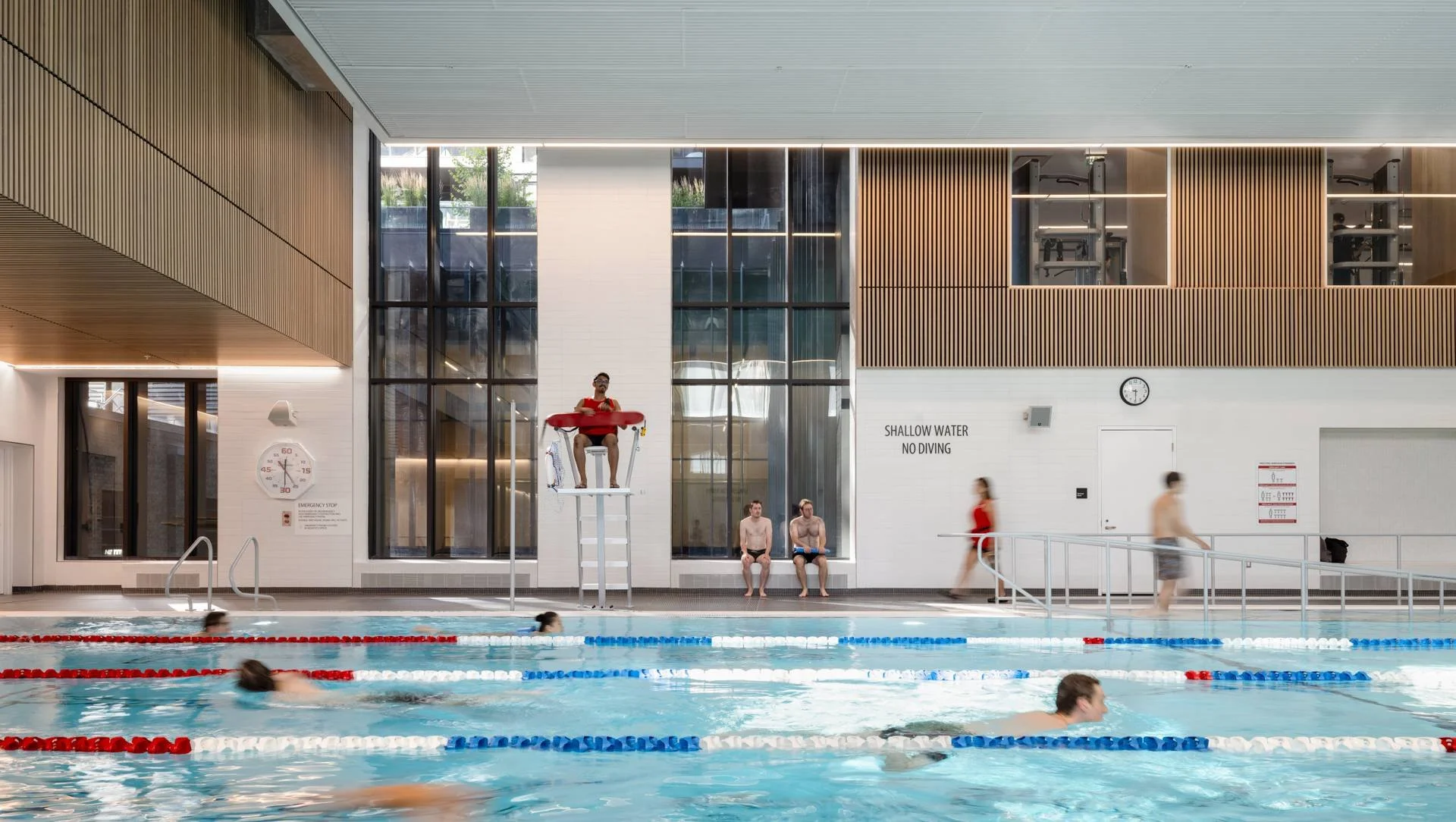 Indoor swimming pool area with people swimming and sitting poolside. A lifeguard in red sits on a high chair. Two people, possibly swimmers, are sitting on the bench. Signs indicate shallow water and no diving. Modern architectural design with wooden panels and large windows.