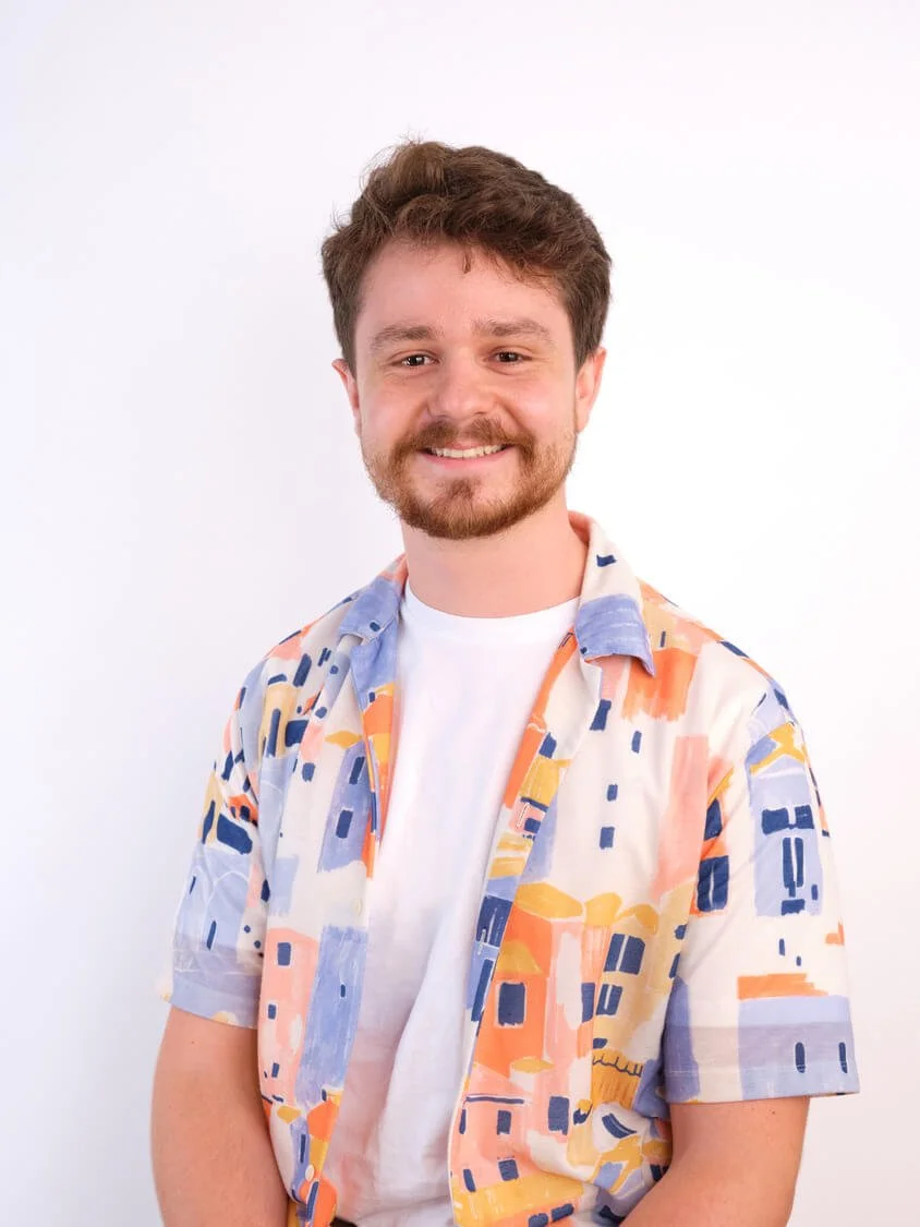 Headshot of a young man with brown hair, a beard, and a mustache smiling at the camera. He is wearing a white t-shirt and an open short-sleeved button-up shirt with a colourful abstract pattern. The background is plain white.