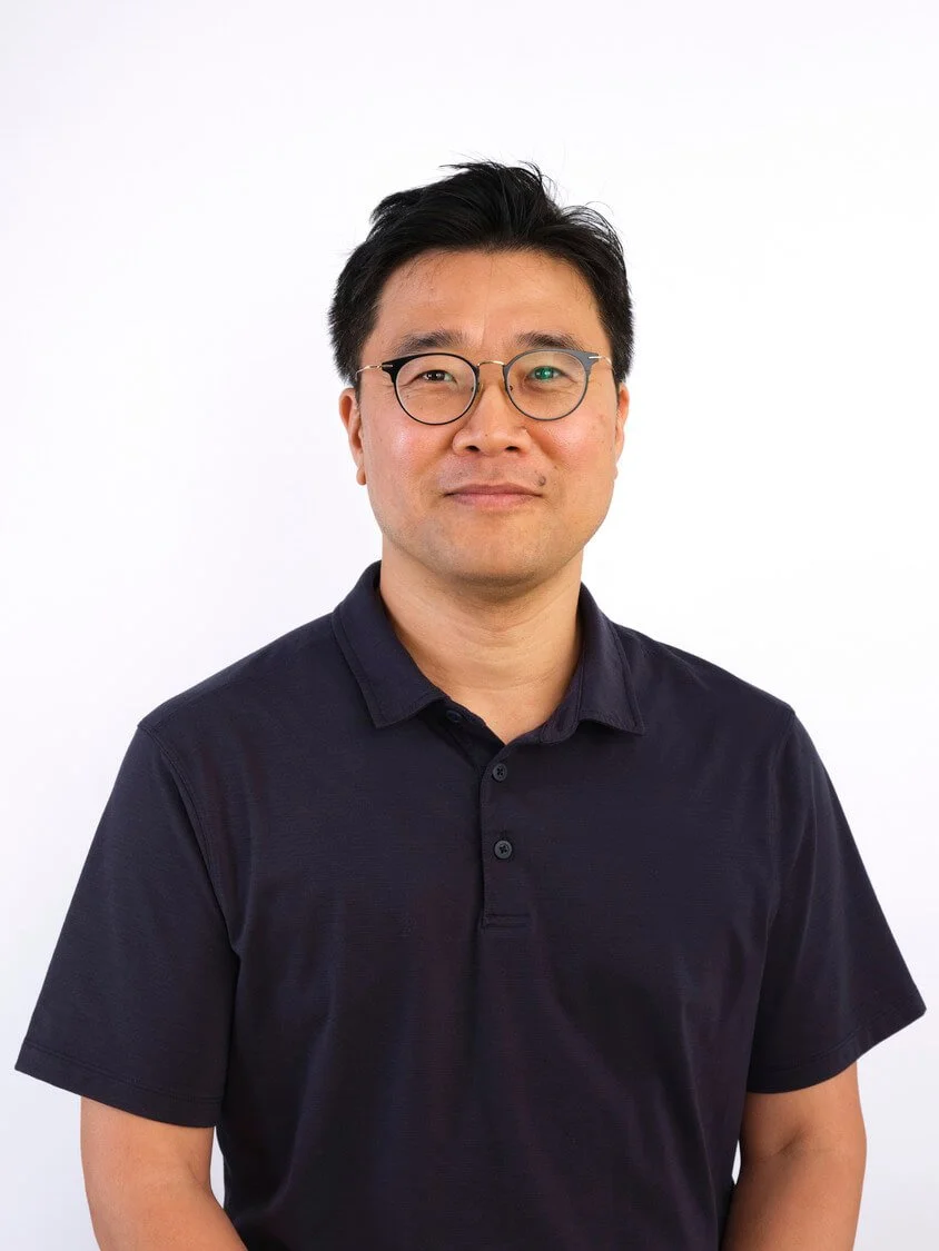 Headshot of a man with short black hair, glasses, and a slight smile, wearing a dark polo shirt, standing against a white background.