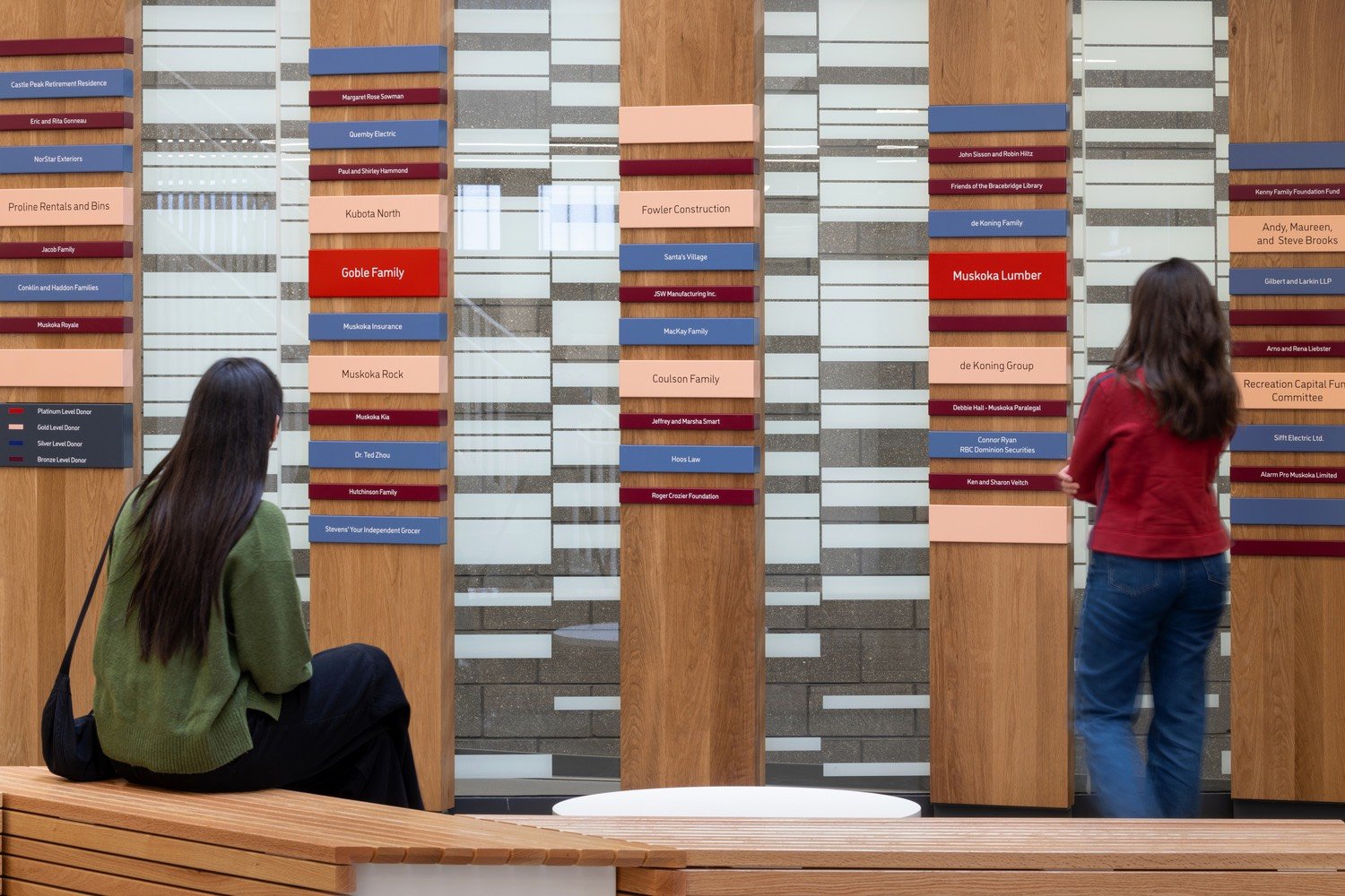 Two women looking at a large donor wall, displaying boards with multiple name plaques in a lobby.