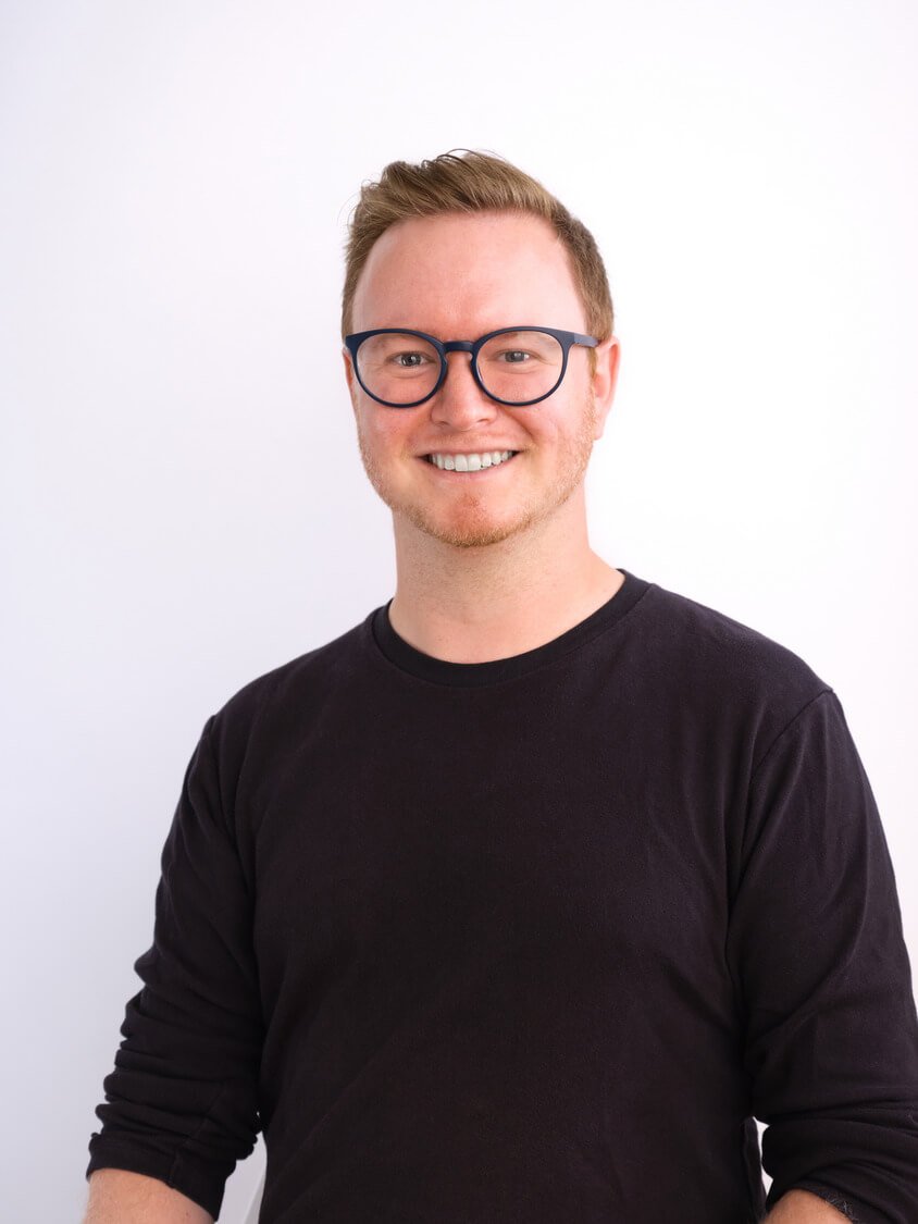 Headshot of a young man, wearing glasses with dark frames, a black long-sleeve shirt, smiling, with short, light brown hair, standing against a plain white background.