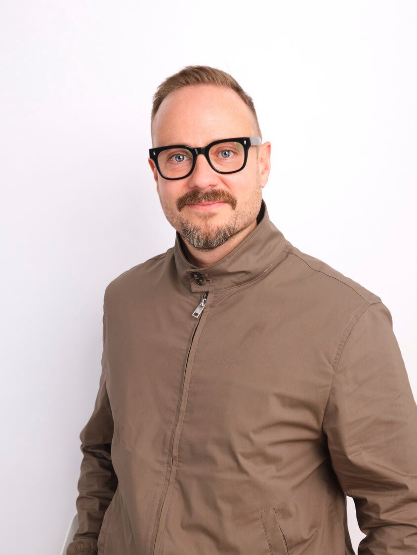 A man with short hair, glasses, and a beard wearing a beige jacket against a white background.