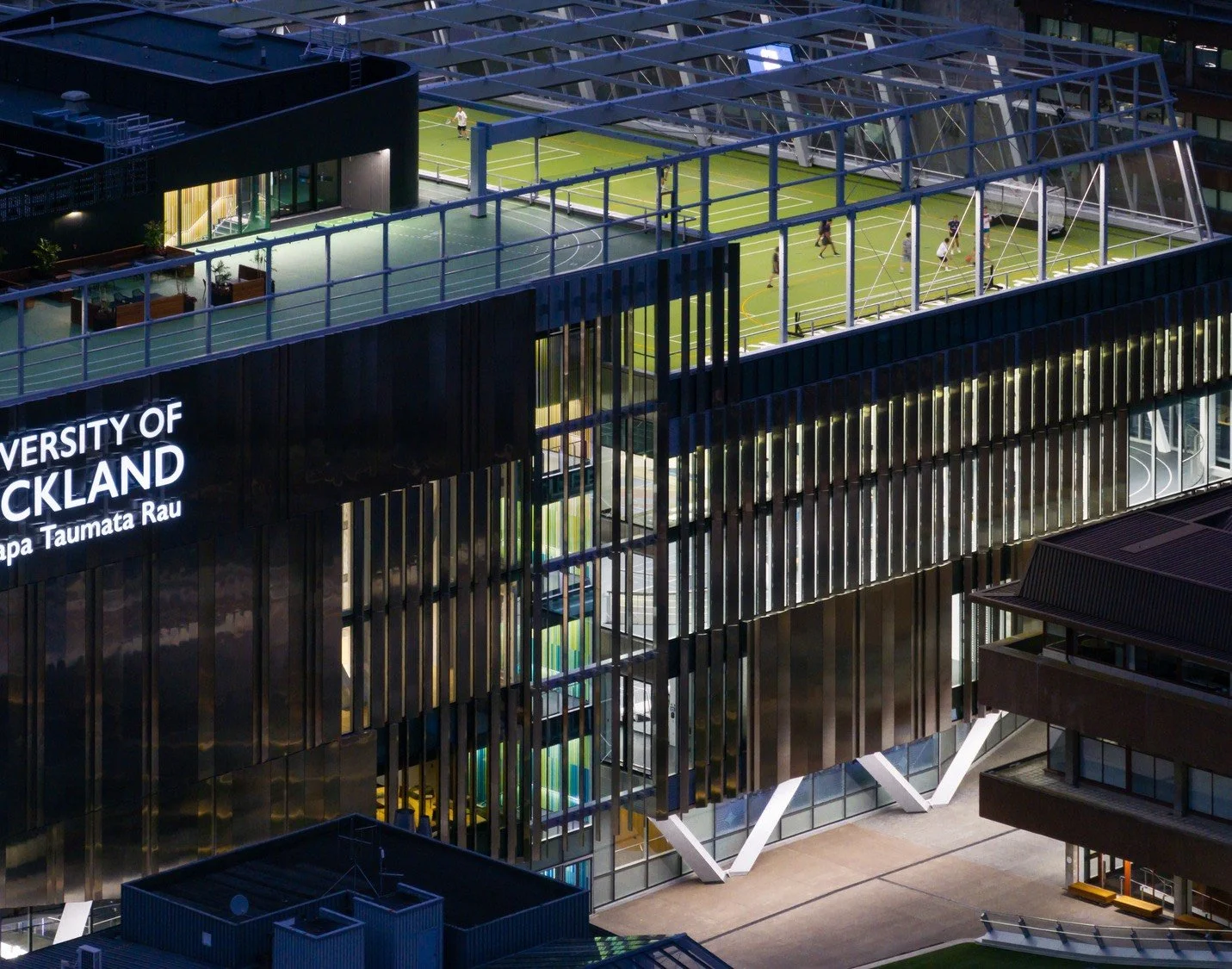 Nighttime view of the University of Auckland campus, featuring a modern building with a rooftop sports area including tennis courts and a soccer field, illuminated and visible through glass windows.