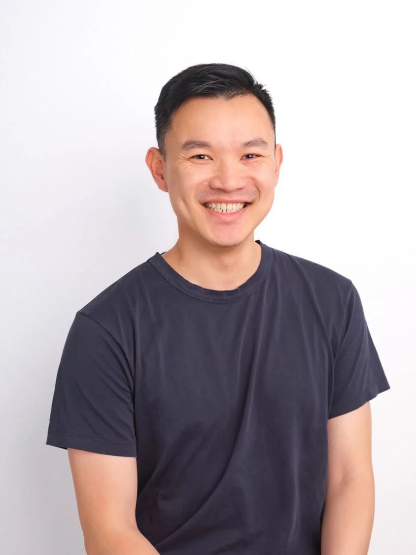 Headshot of a smiling man with short black hair wearing a black t-shirt, standing against a white background.