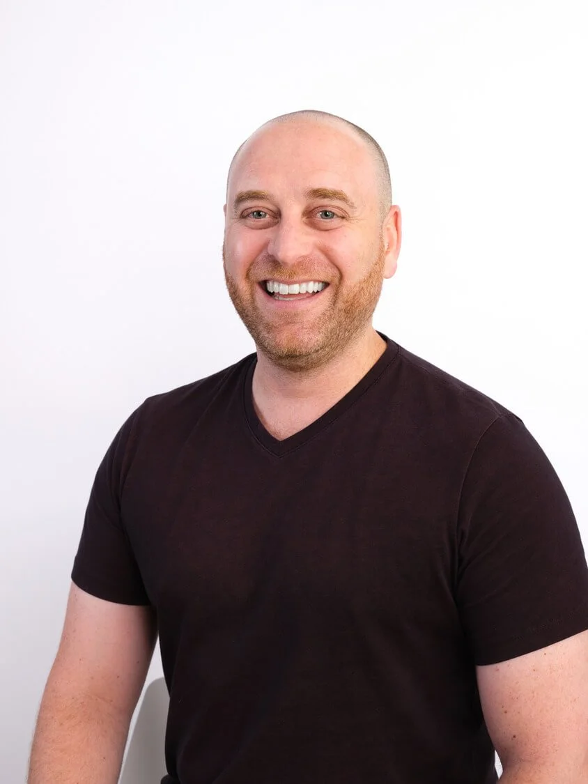 Headshot of a smiling man with a shaved head and beard, wearing a black V-neck T-shirt, sitting against a white background.