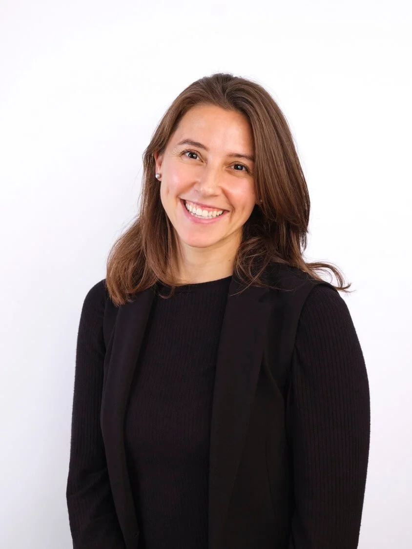 Headshot of a woman with shoulder-length brunette hair wearing a black blazer and black top, smiling against a white background.