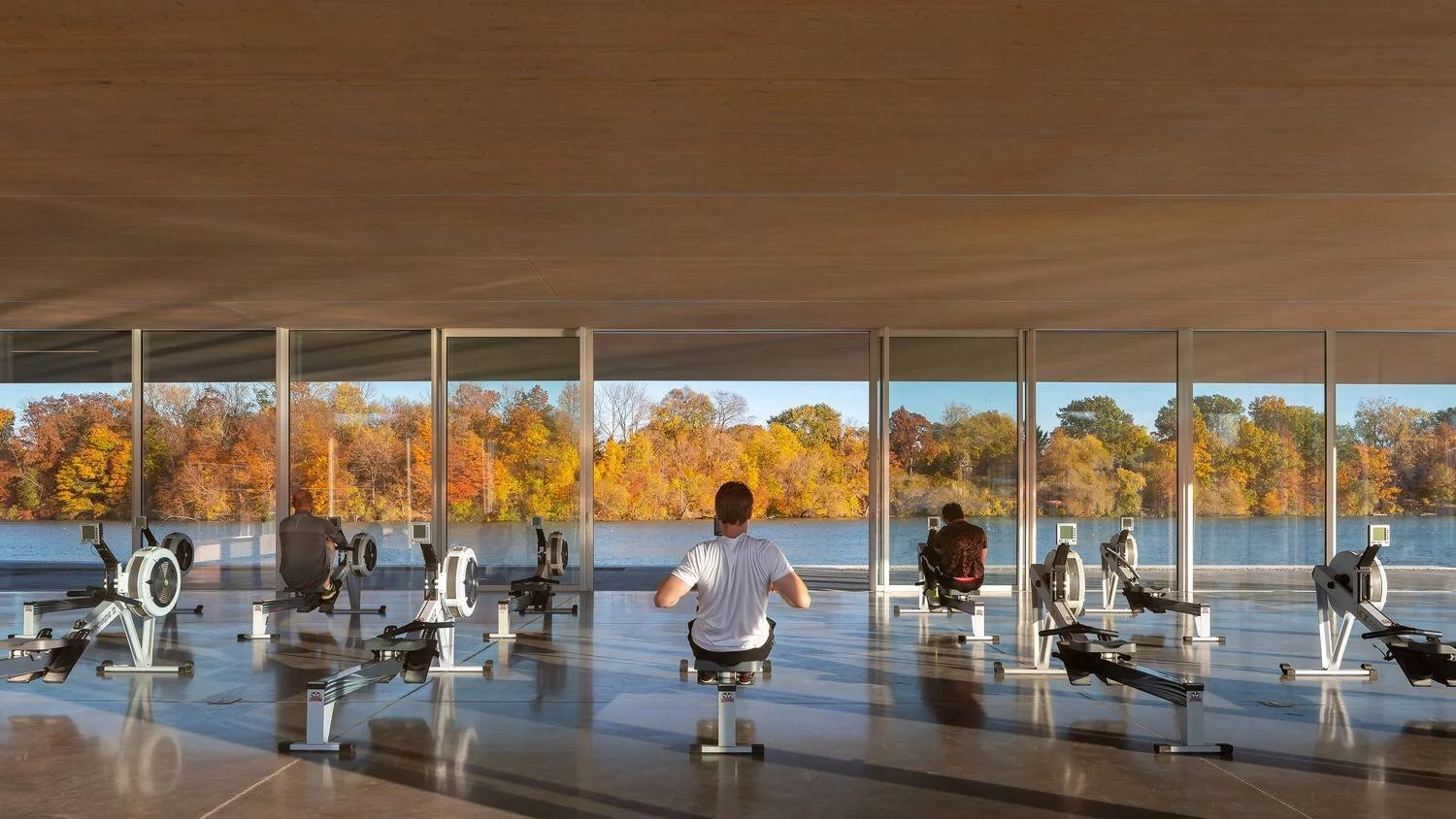 People sitting on rowing machines facing large windows overlooking a lake with colorful autumn trees.