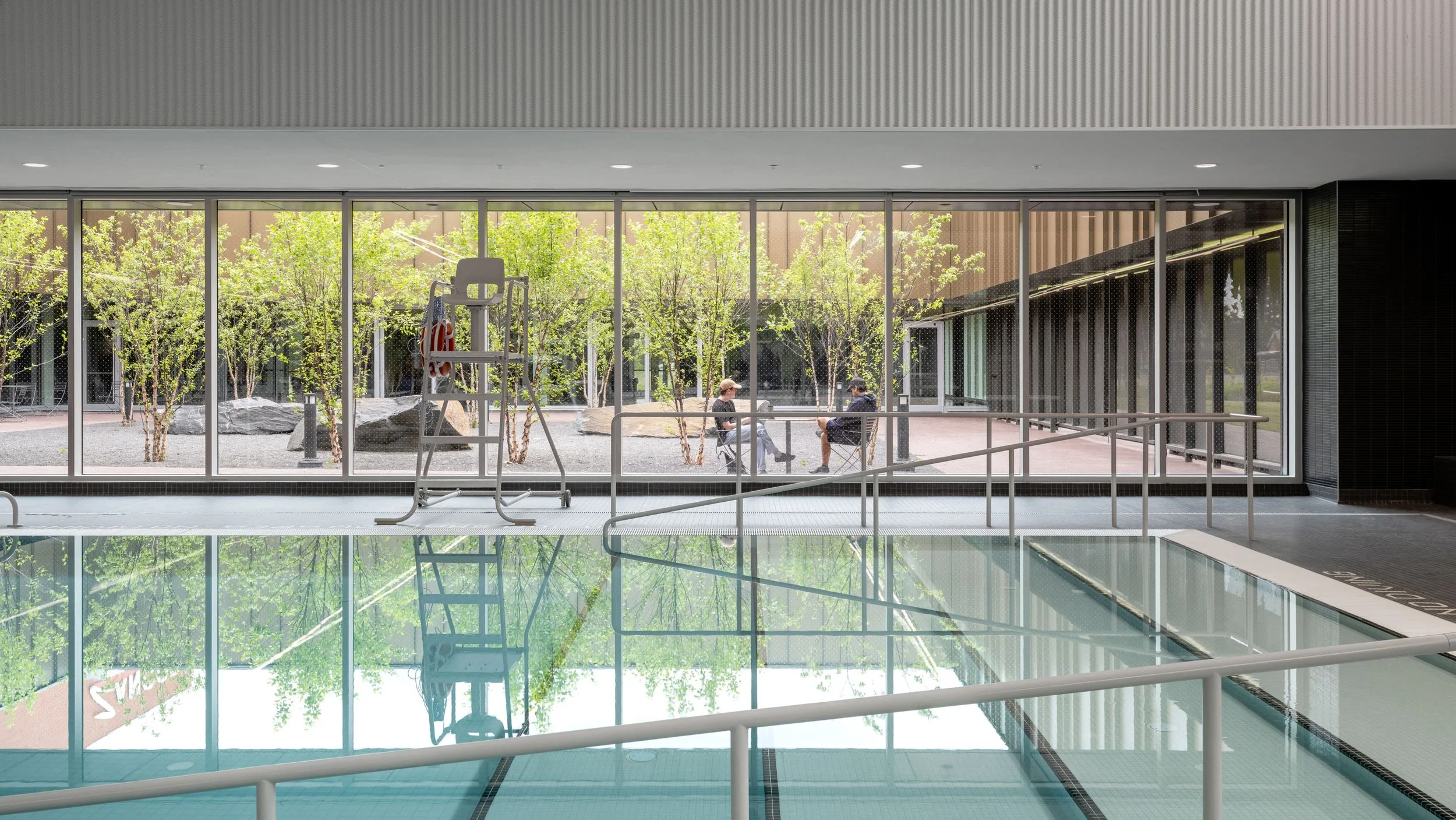 Indoor swimming pool area with a ladder and a railing, overlooking an outdoor patio with trees and two people sitting and talking.