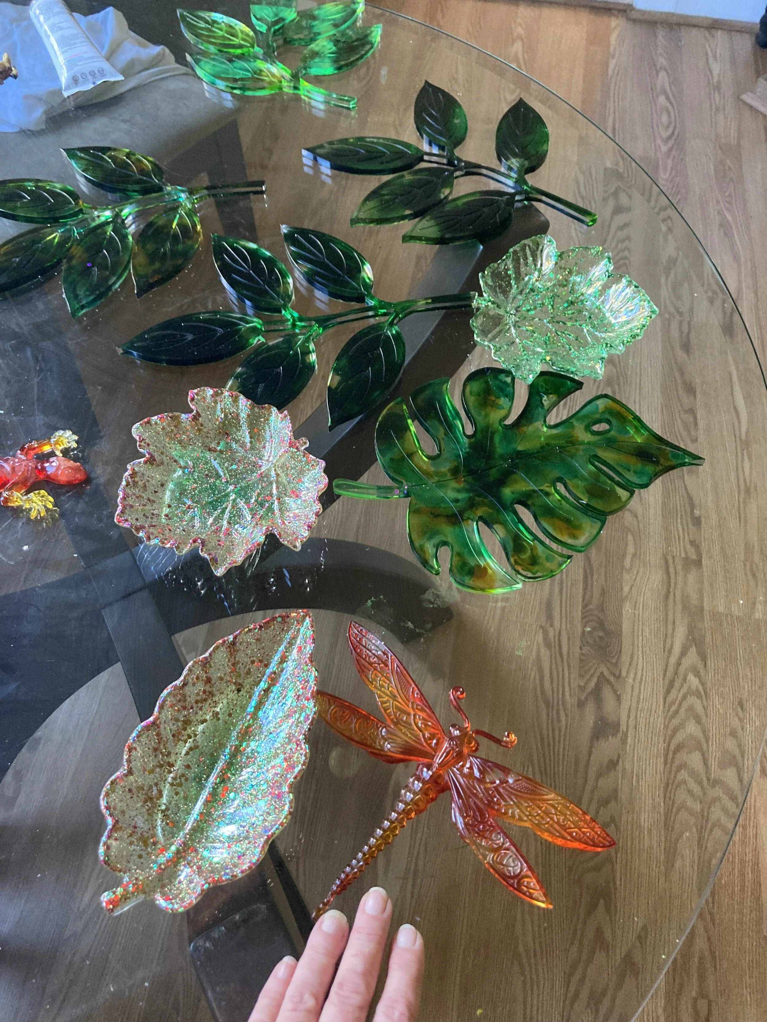 Resin decorative items on a glass table, including green leaves, colorful glittery leaves, a large green monstera leaf, and an orange dragonfly.