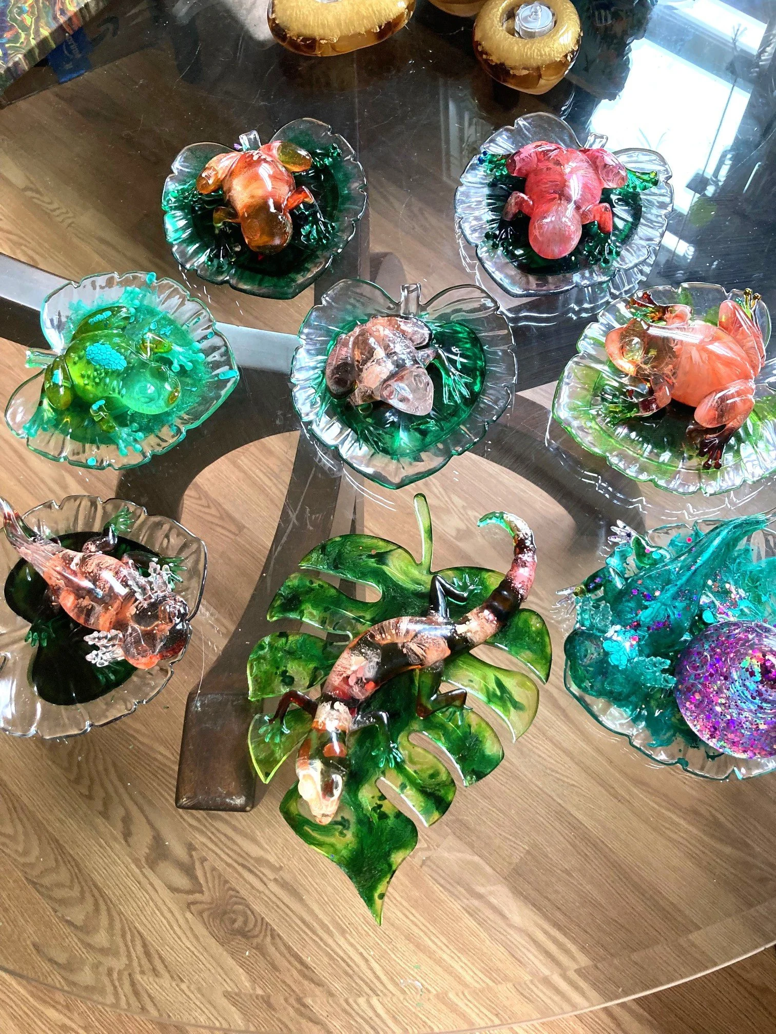Resin art sculptures in the form of animals and insects displayed on glass plates and a glass table. Items include lizards, frogs, crabs,  and other sea creatures, all crafted with vibrant colors and intricate details.