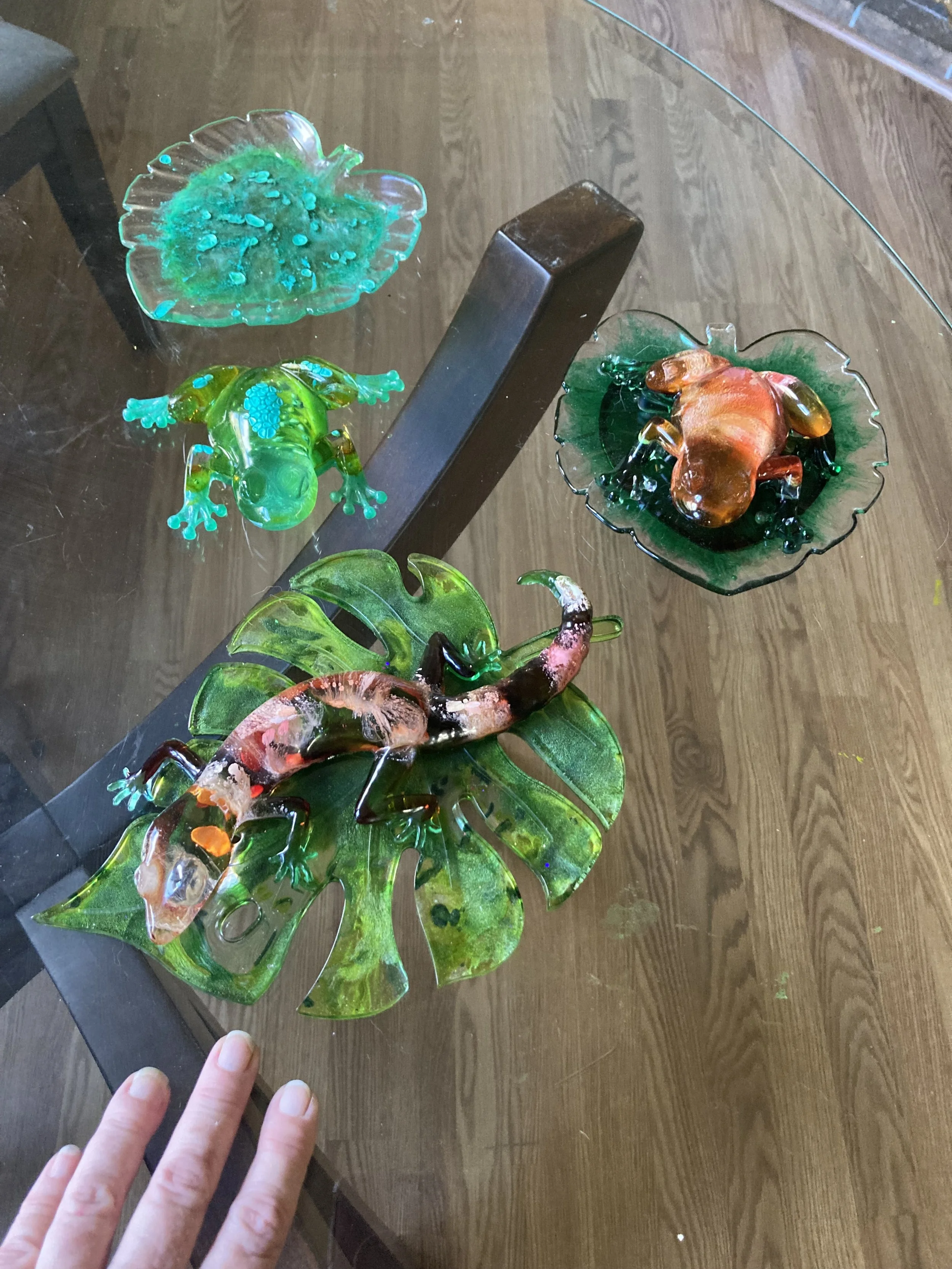 Colorful Resin animal sculptures, including a frog, a lizard, and a crab, displayed on a glass table with a wooden floor underneath.
