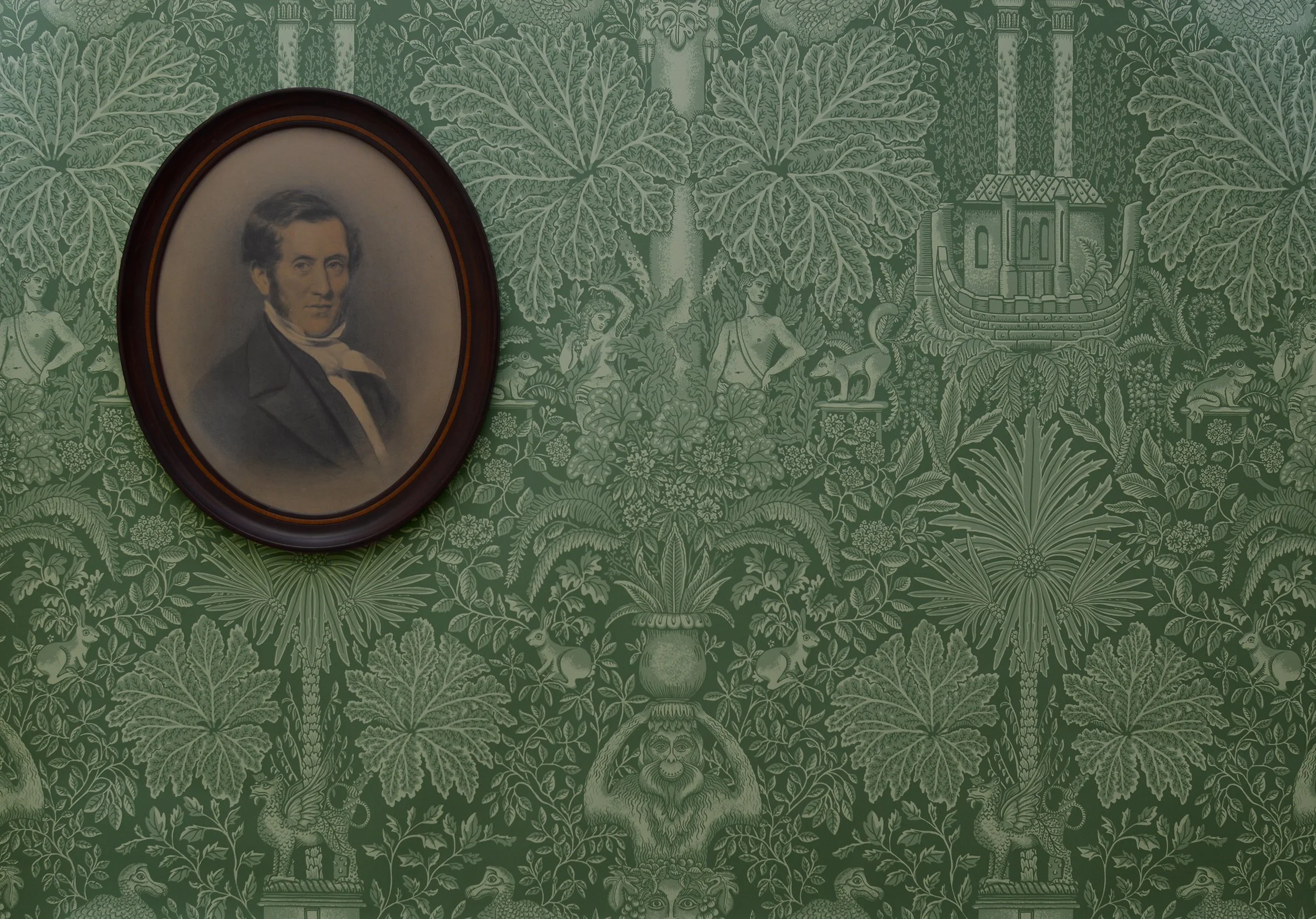 A portrait of a man in 19th centruy clothes hangs on a wall covered in green wallpaper patterned with plants and sculptures in a garden