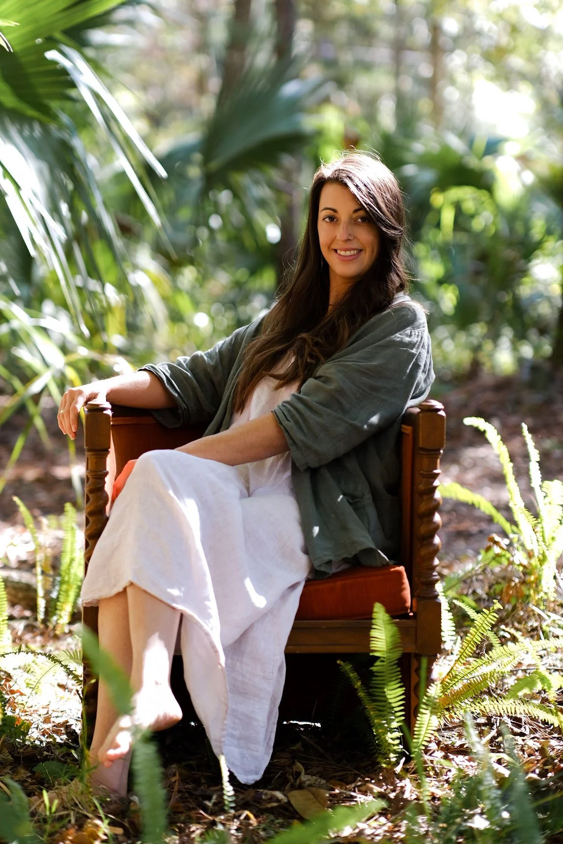 A woman with long dark hair sitting on a wooden chair outdoors in a forest with sunlight filtering through the trees, wearing a light-colored dress and a green jacket, smiling at the camera.