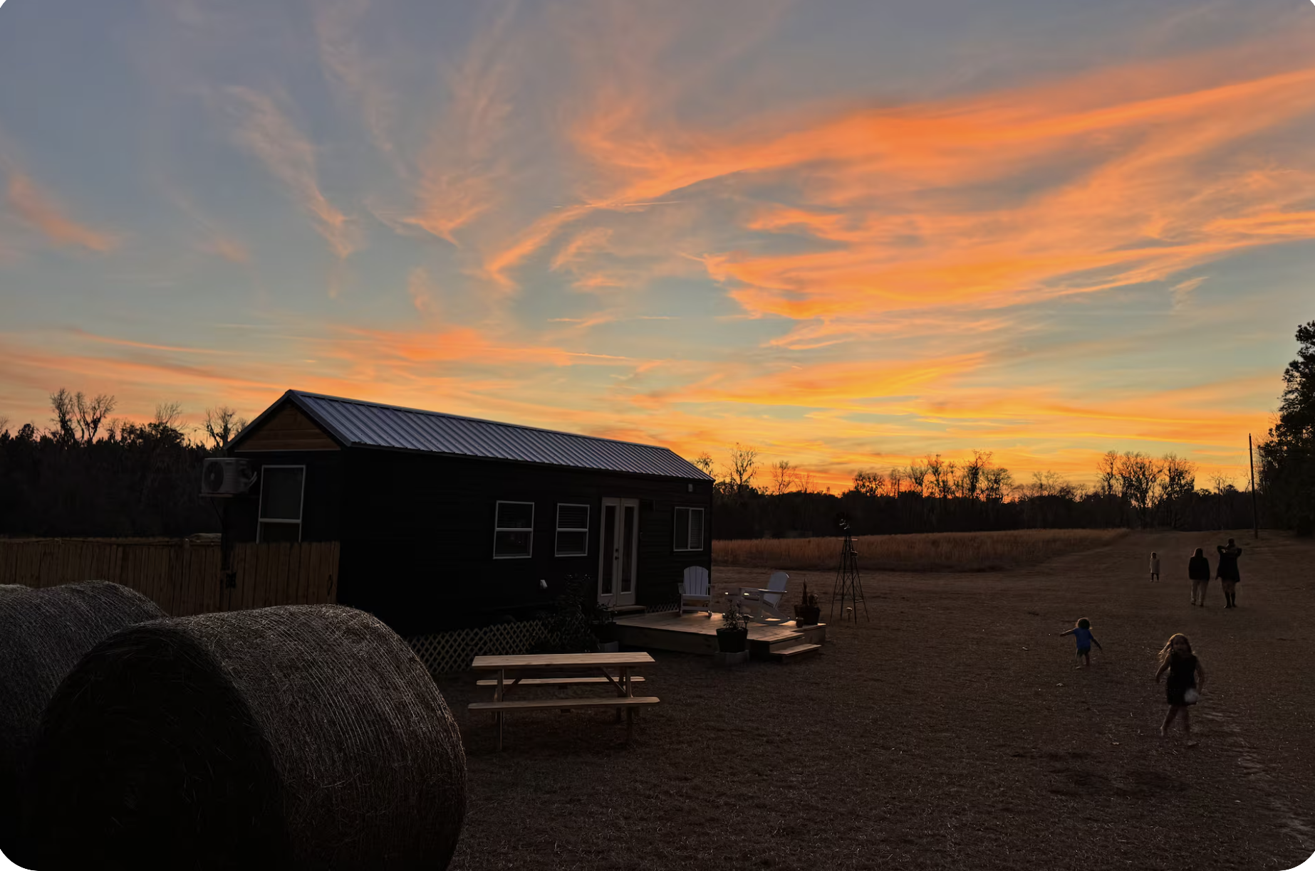 A rural scene during sunset with a house, hay bales, and people, including children, outdoors.