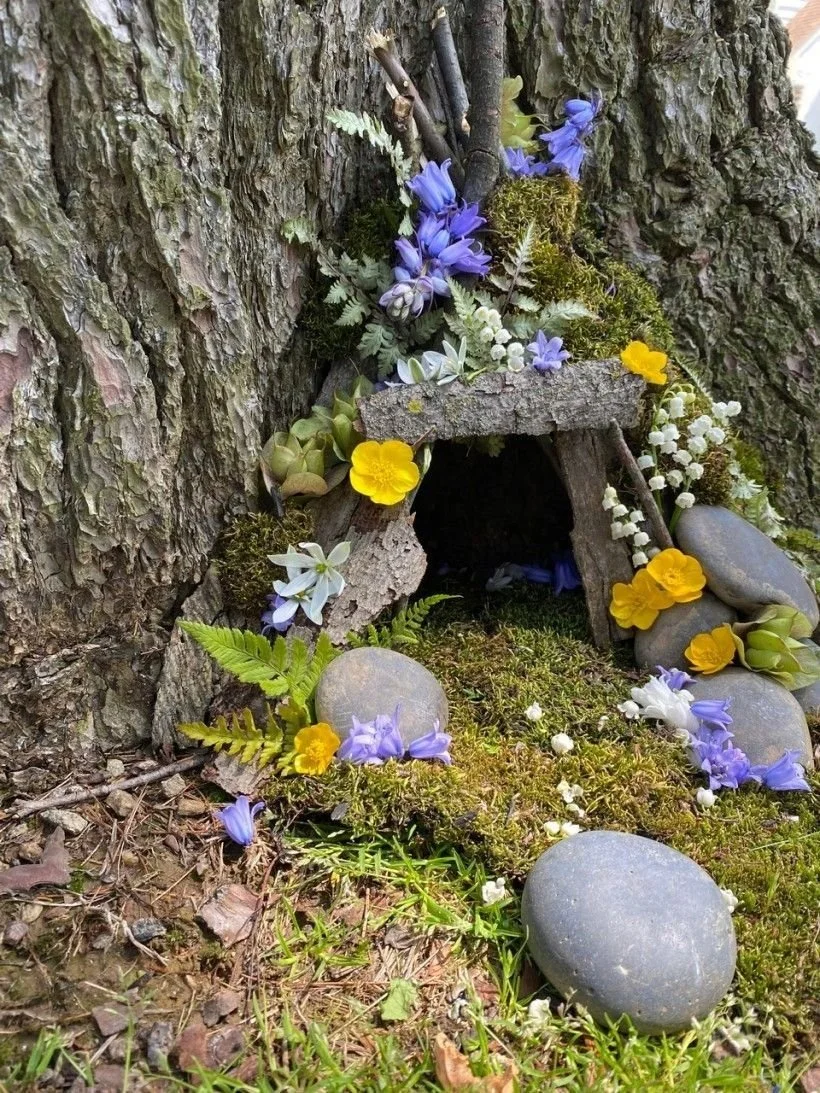 A small fairy house for garden fairies or elves, made of natural materials like bark, sticks, moss, and stones, decorated with purple, yellow, white flowers, and green foliage, placed against a tree trunk.