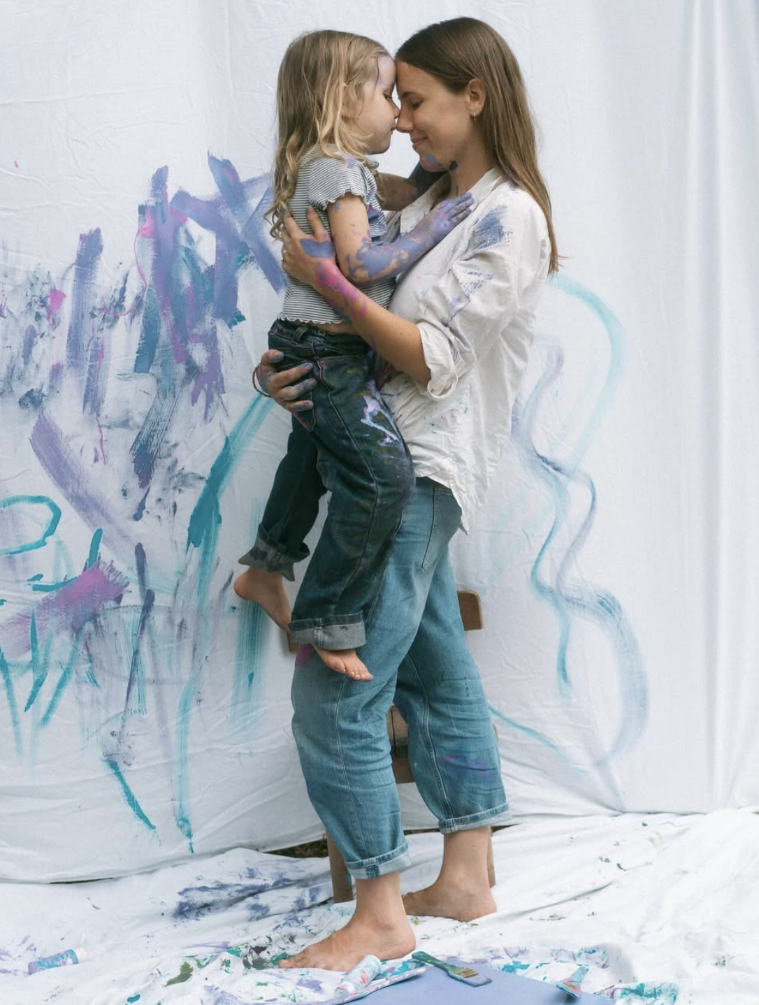 A woman and a young girl touching foreheads while smiling, covered in colorful paint, standing against a painted backdrop.