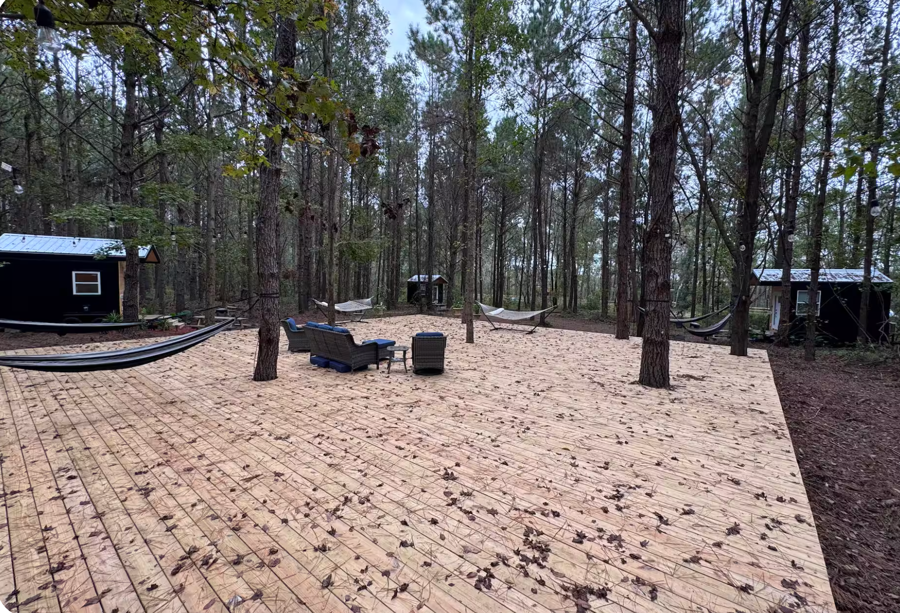 A newly built wooden deck in a forest, with outdoor furniture, hammocks, and small structures, surrounded by tall trees and fallen leaves.