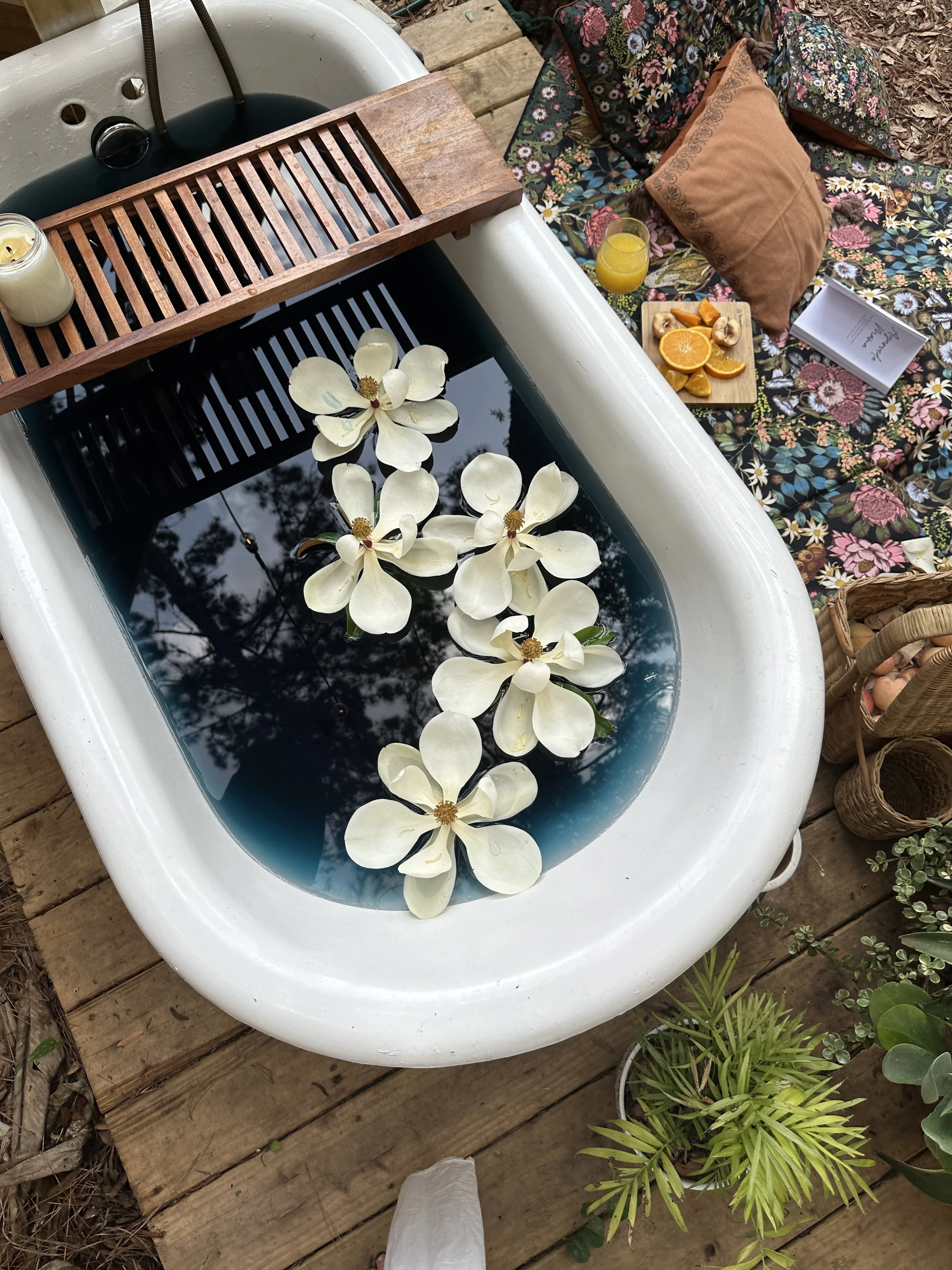 A vintage bathtub filled with water and floating white magnolia flowers. Beside the tub, there is a wooden tray, a candle, a glass of orange juice, a wooden tray with sliced oranges and bananas, a floral patterned blanket with pillows, and some greenery on a wooden deck.