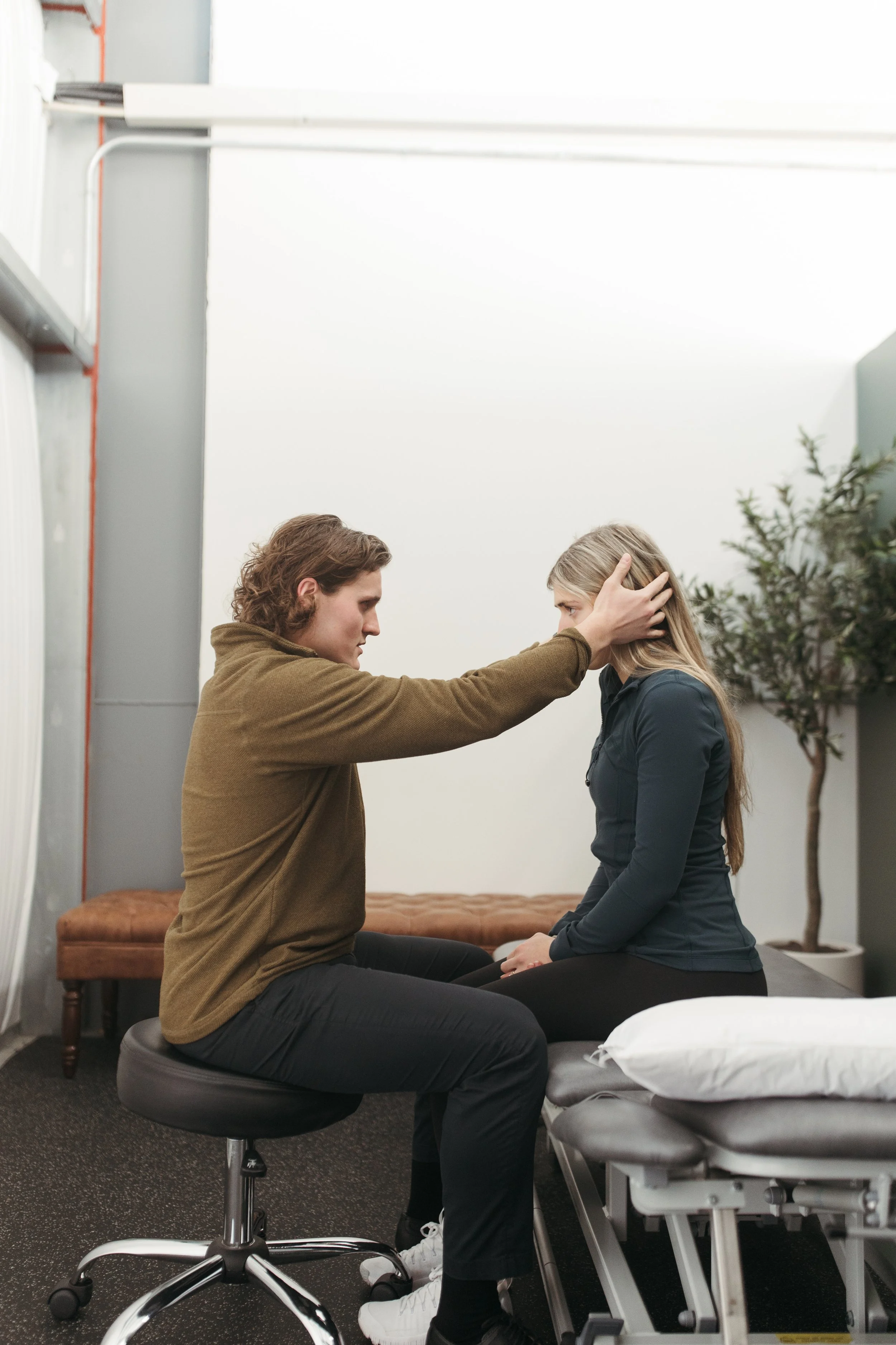 A man kneeling in front of a woman, with his hands on her head, in a therapeutic or counseling setting. The woman is sitting on a table with her eyes closed, and the man appears to be offering comfort or support. They are in a room with a plant and some furniture.