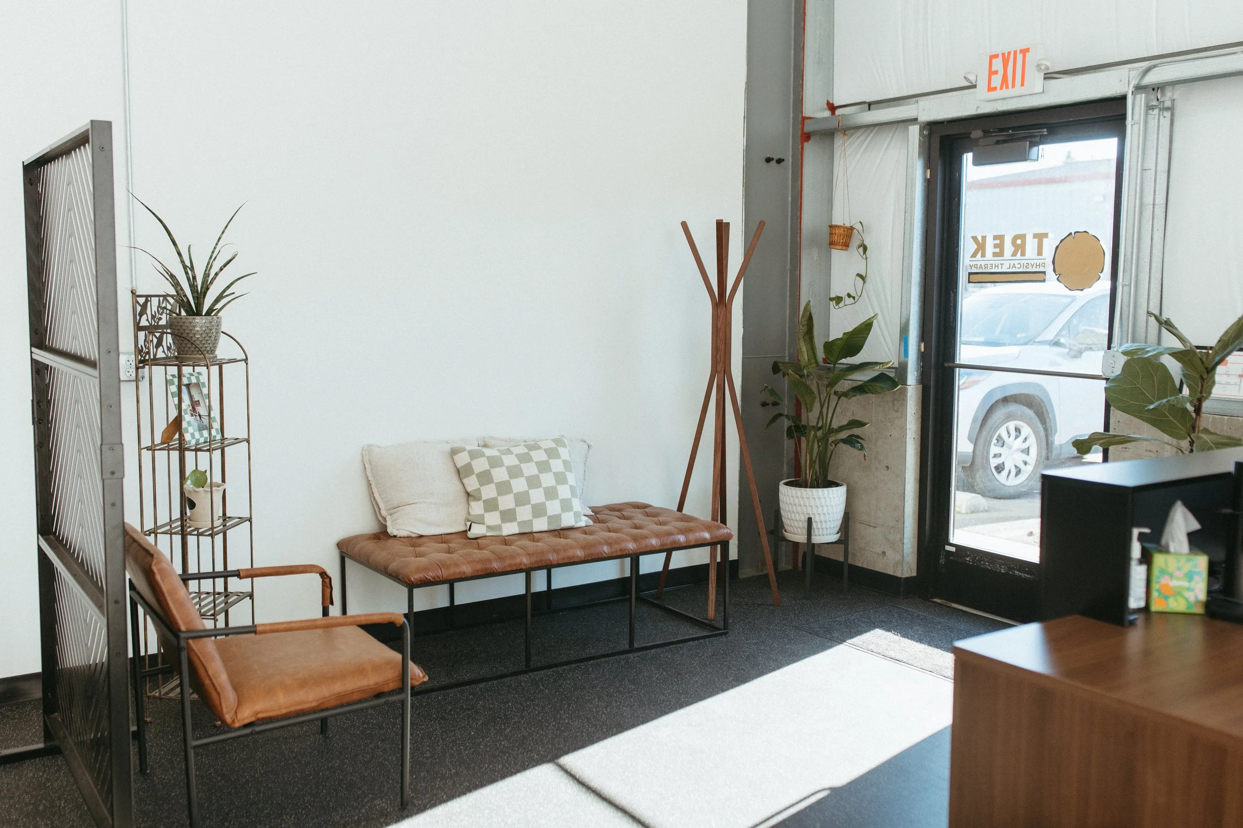 Interior of a waiting area with a brown leather armchair, a tan tufted bench with pillows, potted plants, a coat rack, and a door leading outside with a sign that says "Trek."