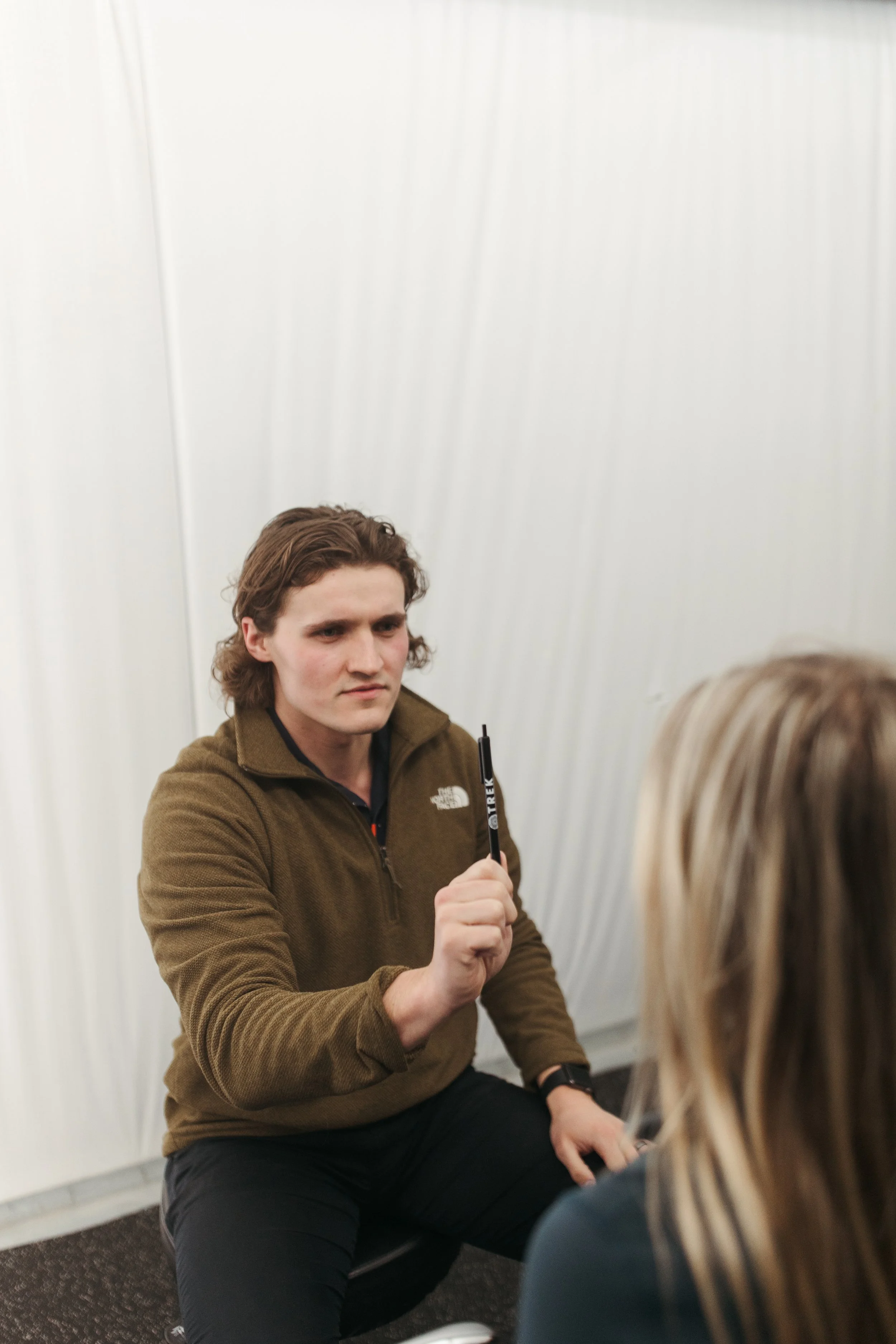 A young man with brown wavy hair wearing a brown North Face jacket kneeling and holding a black marker, speaking to a woman with long blonde hair in front of him, against a plain white wall.