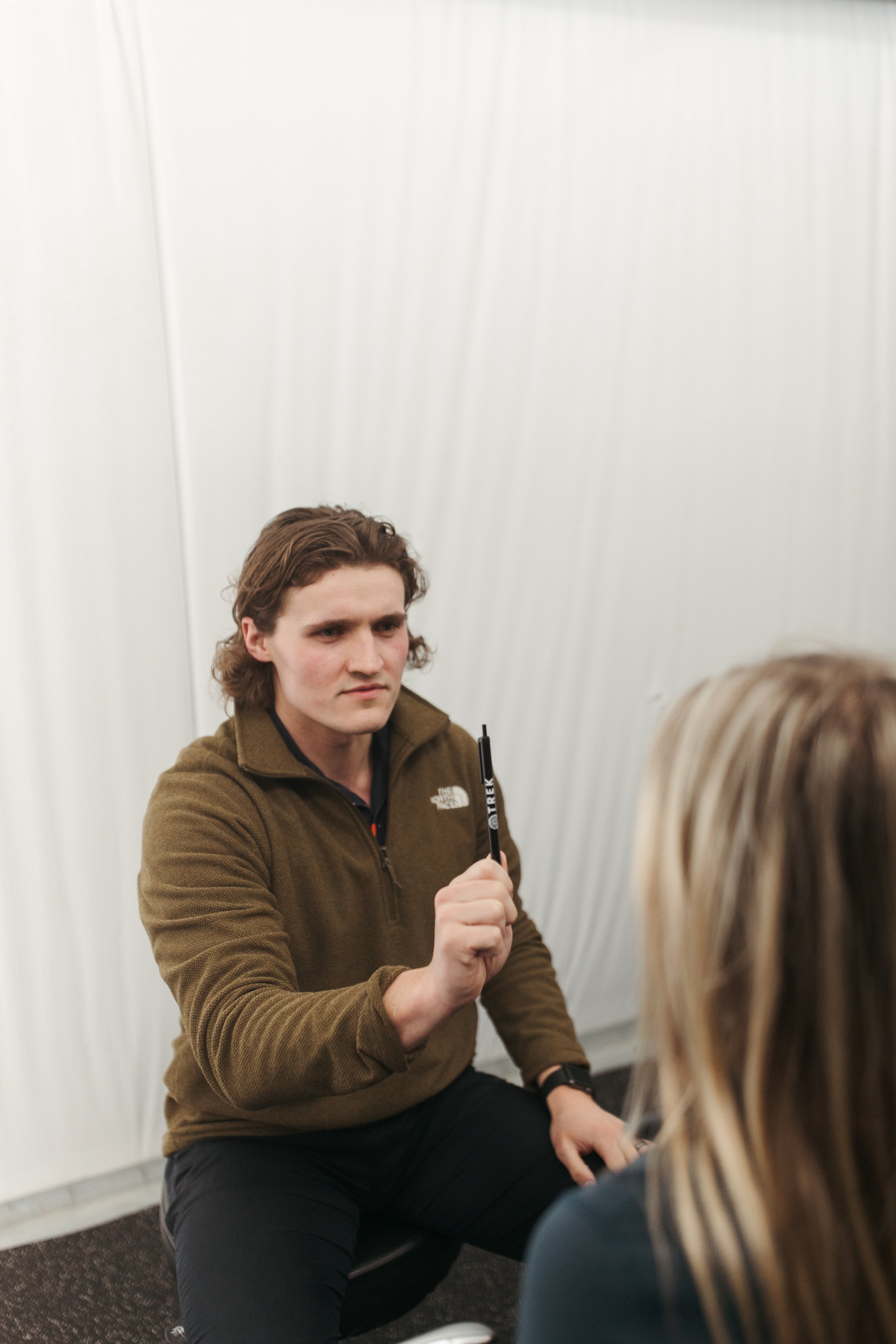 A man with shoulder-length brown hair, wearing a brown North Face jacket, is sitting down and holding a marker while speaking to a person with blonde hair, in an indoor setting with plain white walls.