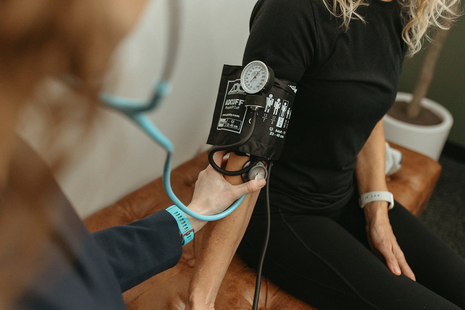 A healthcare professional measures an elderly woman's blood pressure using a sphygmomanometer while she sits on a brown couch in a medical setting. RN provides education and helps coordinate care.