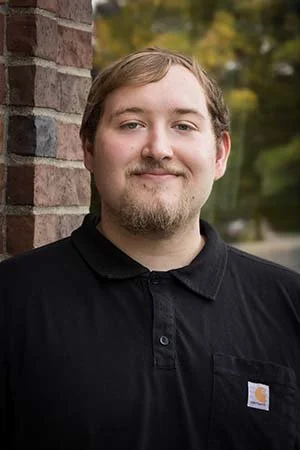 A person wearing a black collared shirt, leaning against a brick wall, outdoors.