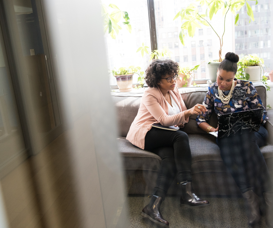 Two women sitting on a gray sofa in an office, reviewing a laptop and a notebook, with large windows and potted plants in the background.