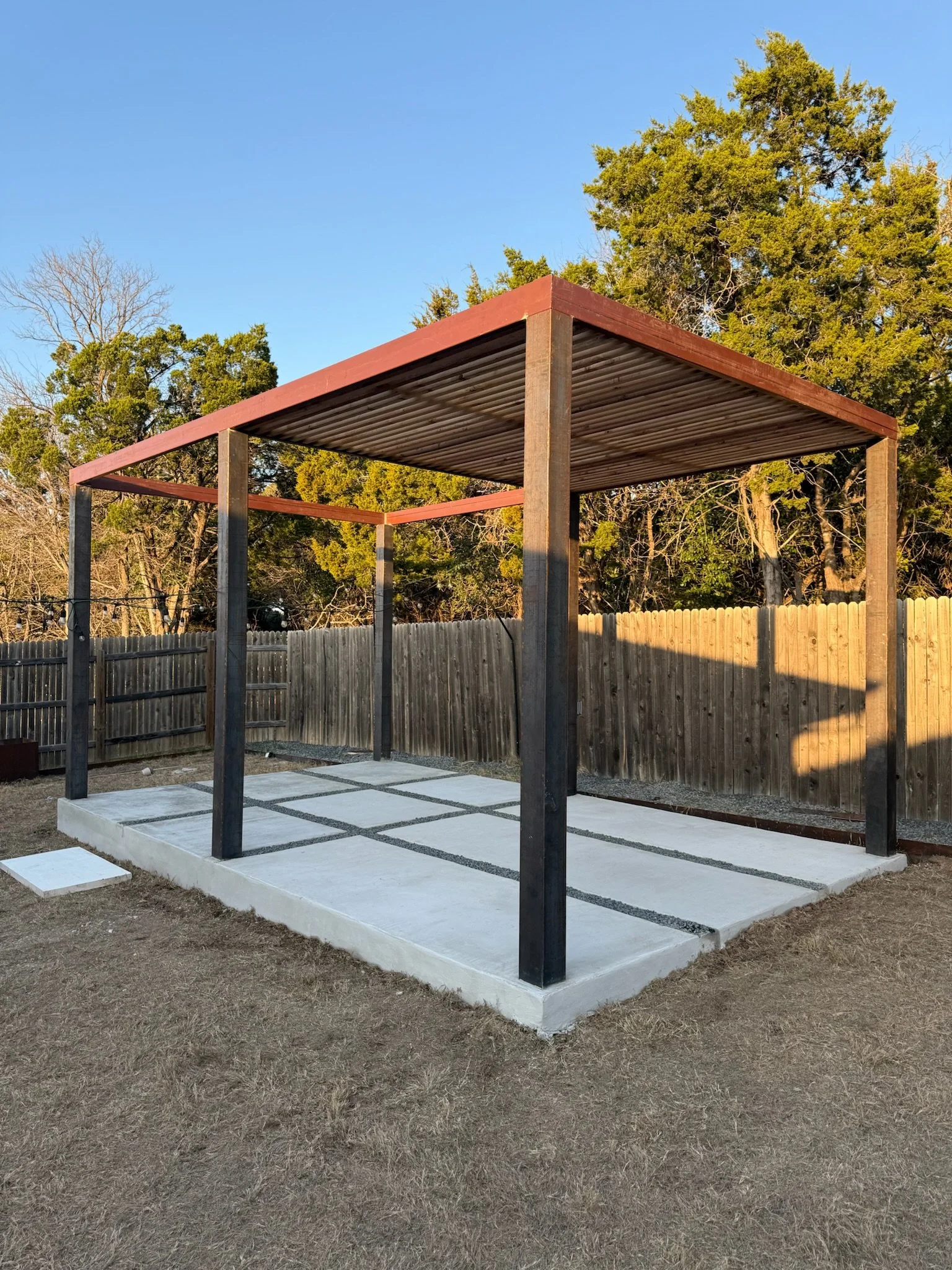 Wooden pergola with a flat roof over a concrete patio, enclosed by a wooden fence, with trees in the background and clear blue sky.