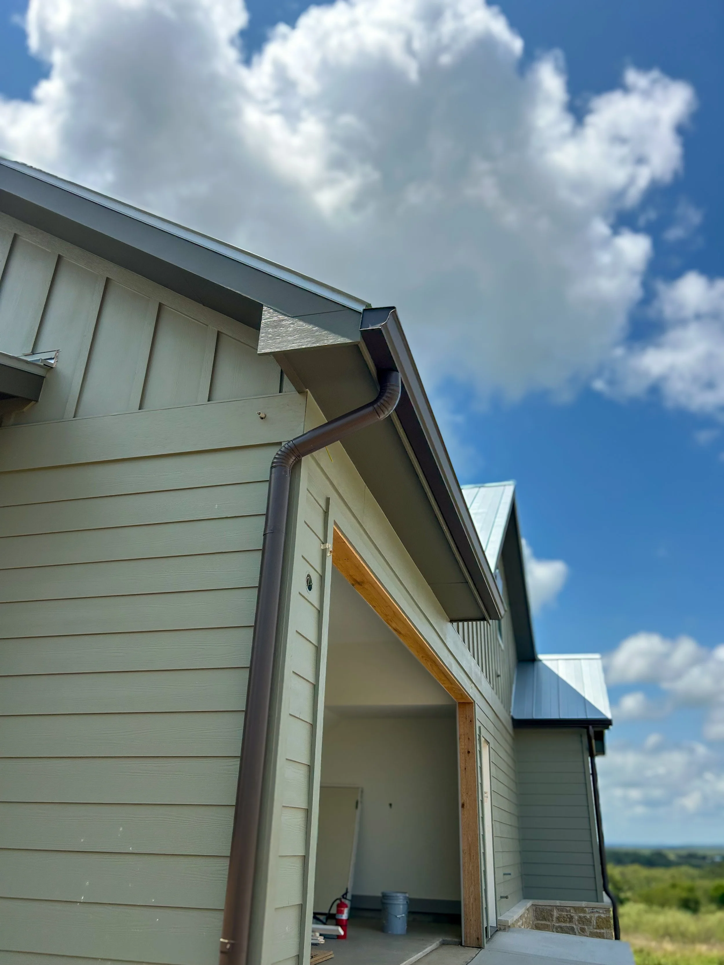 Side view of a house under construction showing light green siding, a roof with metal panels, a black gutter, and an open garage with construction tools inside, set against a blue sky with clouds.