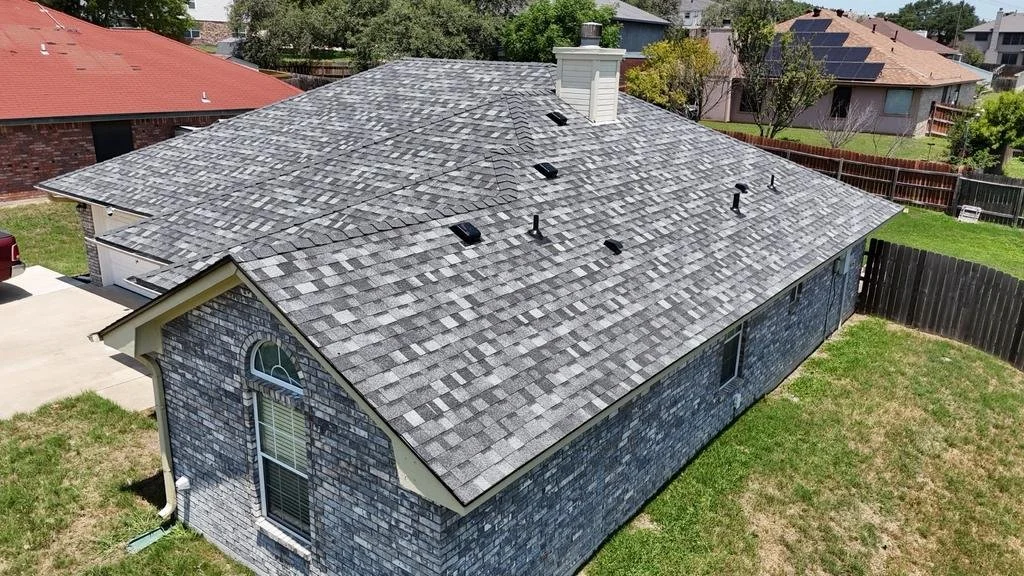 Aerial view of a house with a gray shingle roof, brick exterior walls, a white gutter system, and a backyard surrounded by a wooden fence.