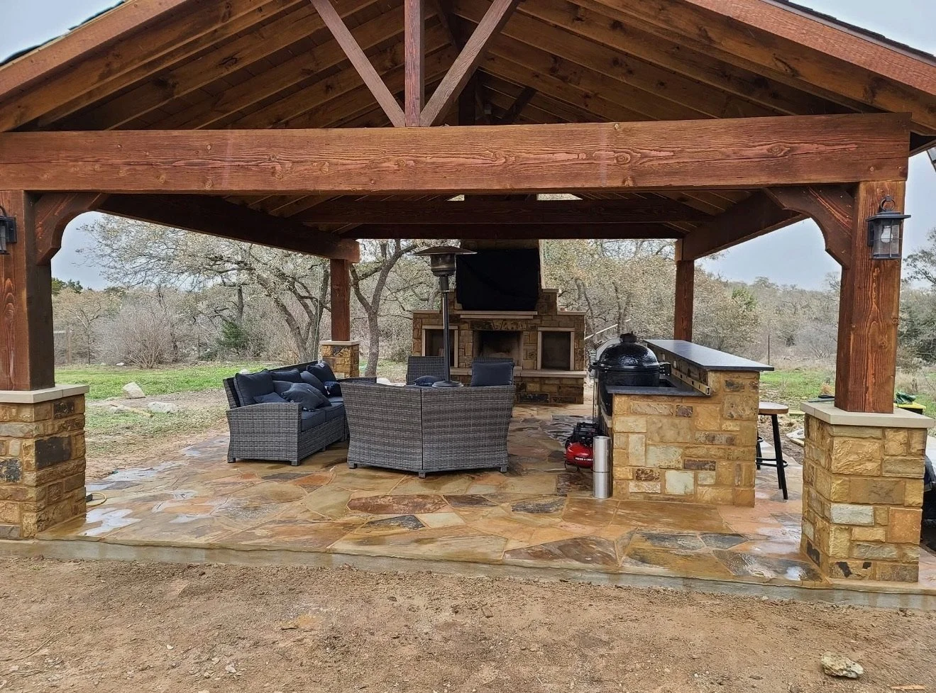 Outdoor covered patio with stone floor, wicker furniture, a TV, fireplace, and a barbecue grill, with a natural landscape and trees in the background.