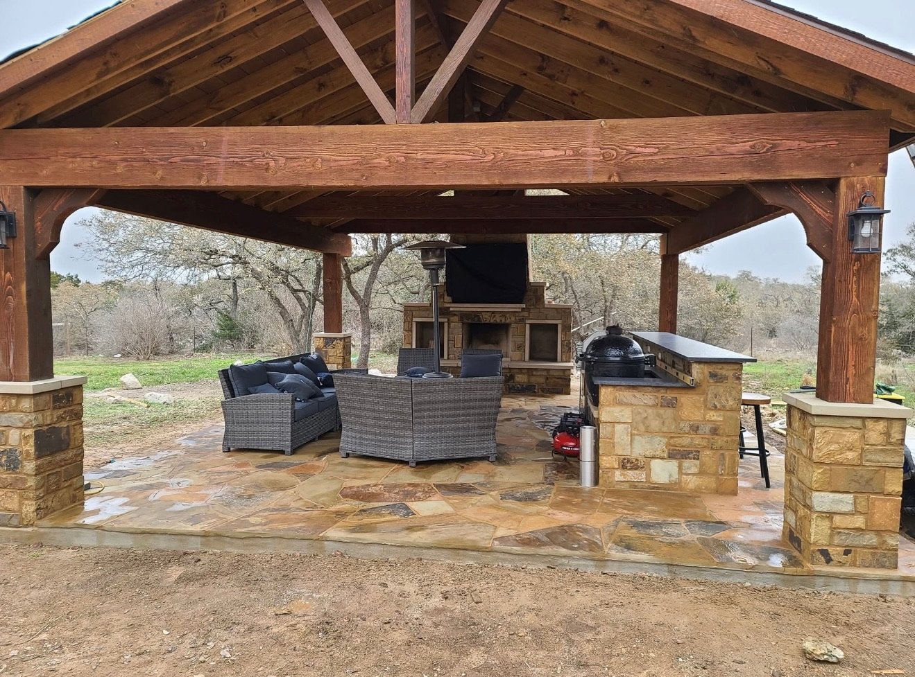 Outdoor pavilion with stone patio, wooden roof, and comfortable seating area featuring wicker furniture. Includes a BBQ area with an outdoor kitchen, surrounded by trees.
