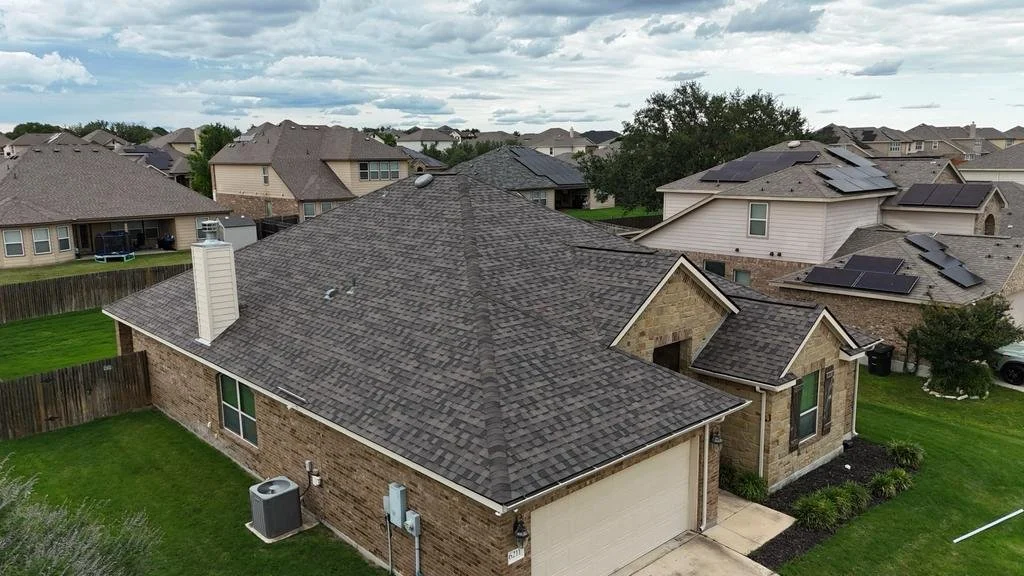 A suburban neighborhood with a view of a house with a gray shingle roof and brick exterior, surrounded by a green lawn and other houses with solar panels on their roofs.