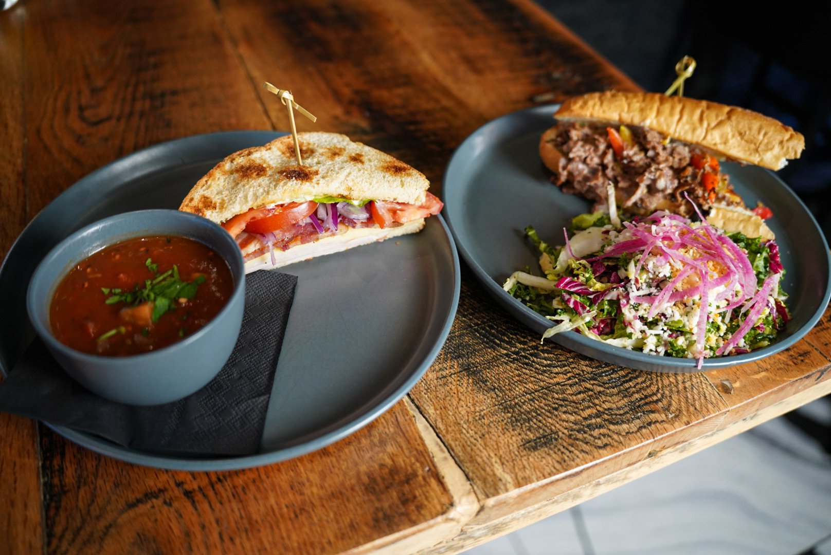 Two plated lunch dishes on a wooden table at South Village Grille, featuring a toasted sandwich with tomato, onion, and soup on one plate, and a chopped steak sandwich with a fresh green salad on the other.