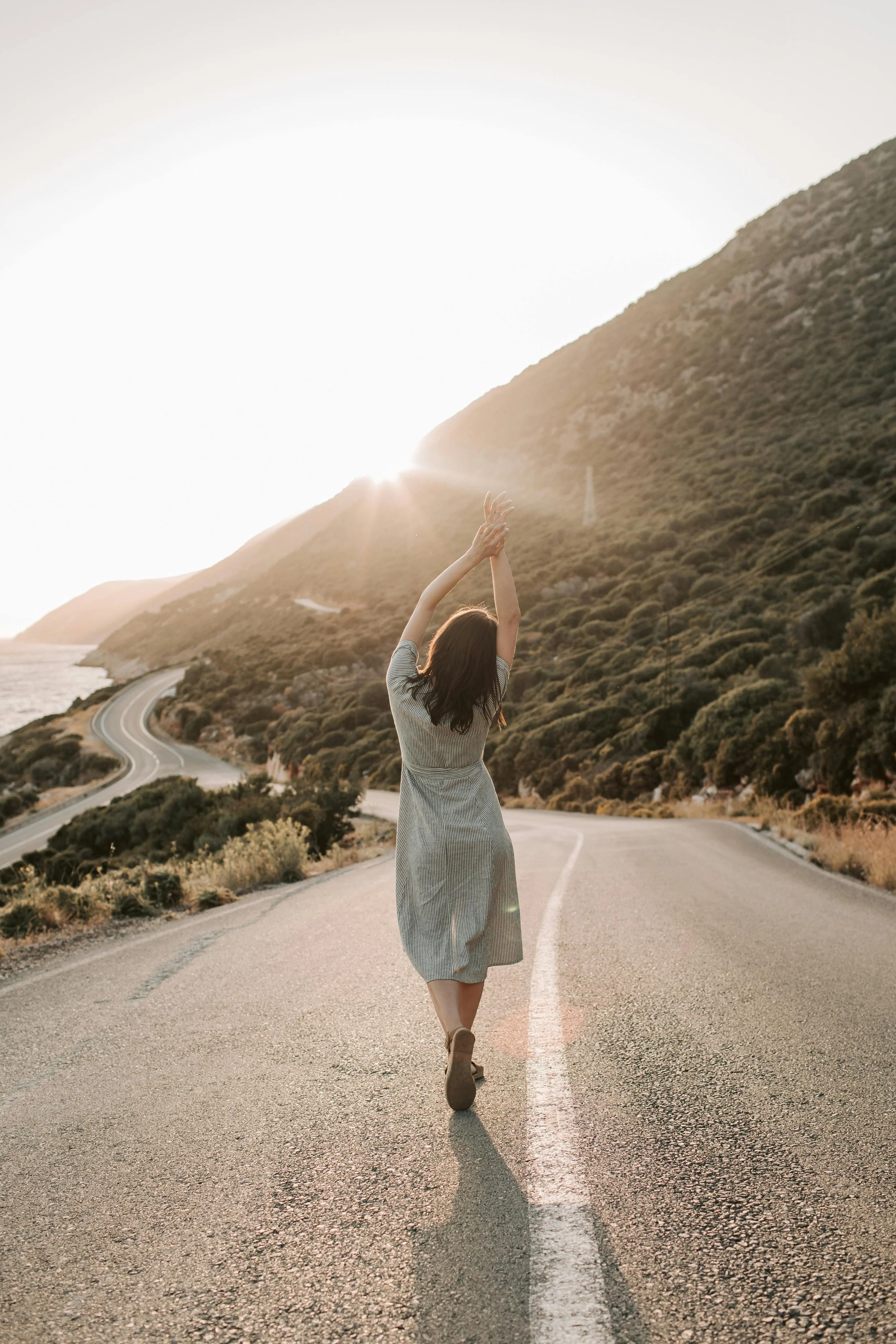 Back view of a woman with dark hair in a green dress standing on a deserted coastal road stretching