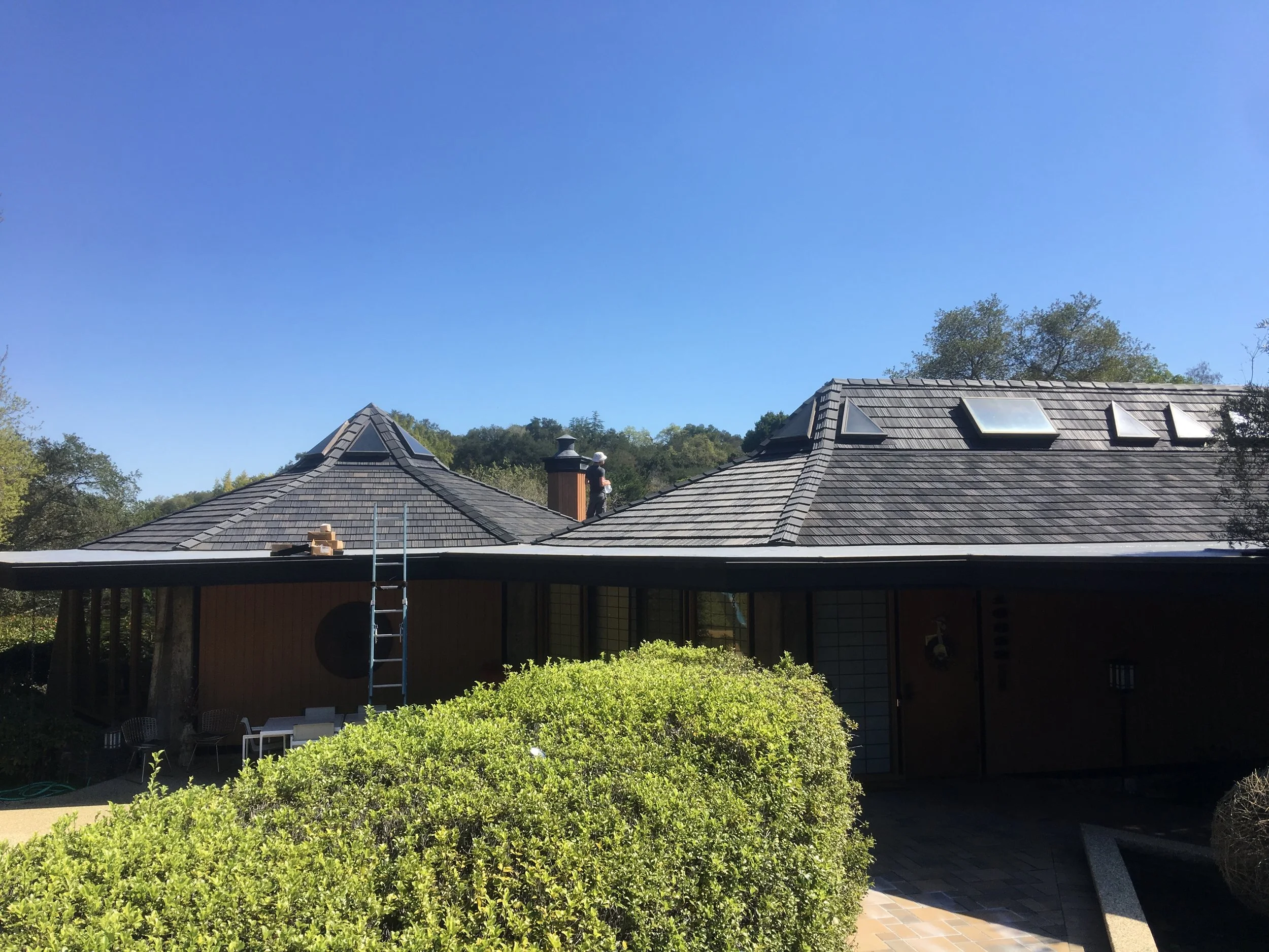 Front view of a house with a shingled roof, several skylights, and a person working on the roof near a chimney under a clear blue sky.