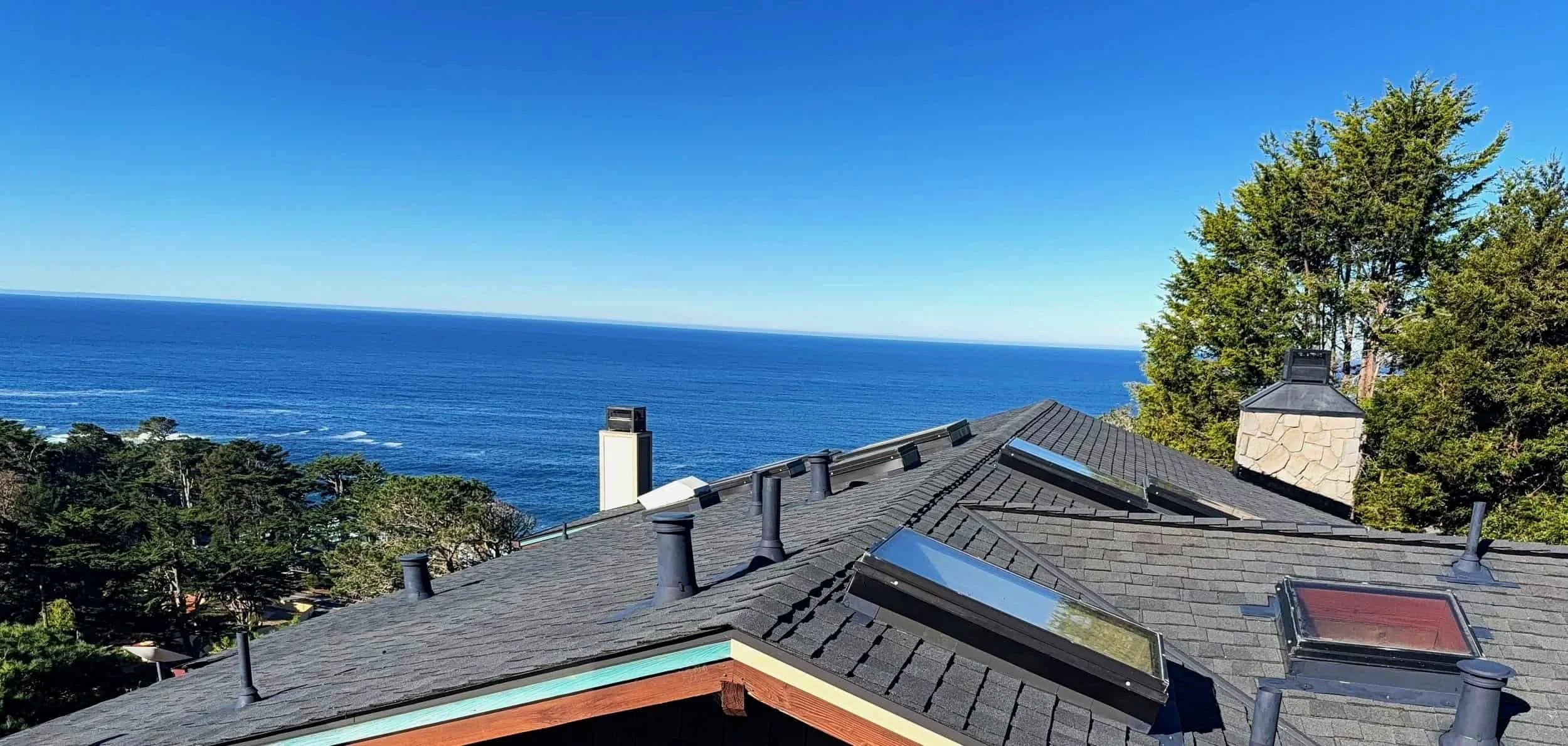 Residential rooftops with vents and skylights overlooking the ocean, surrounded by trees, under a clear blue sky.