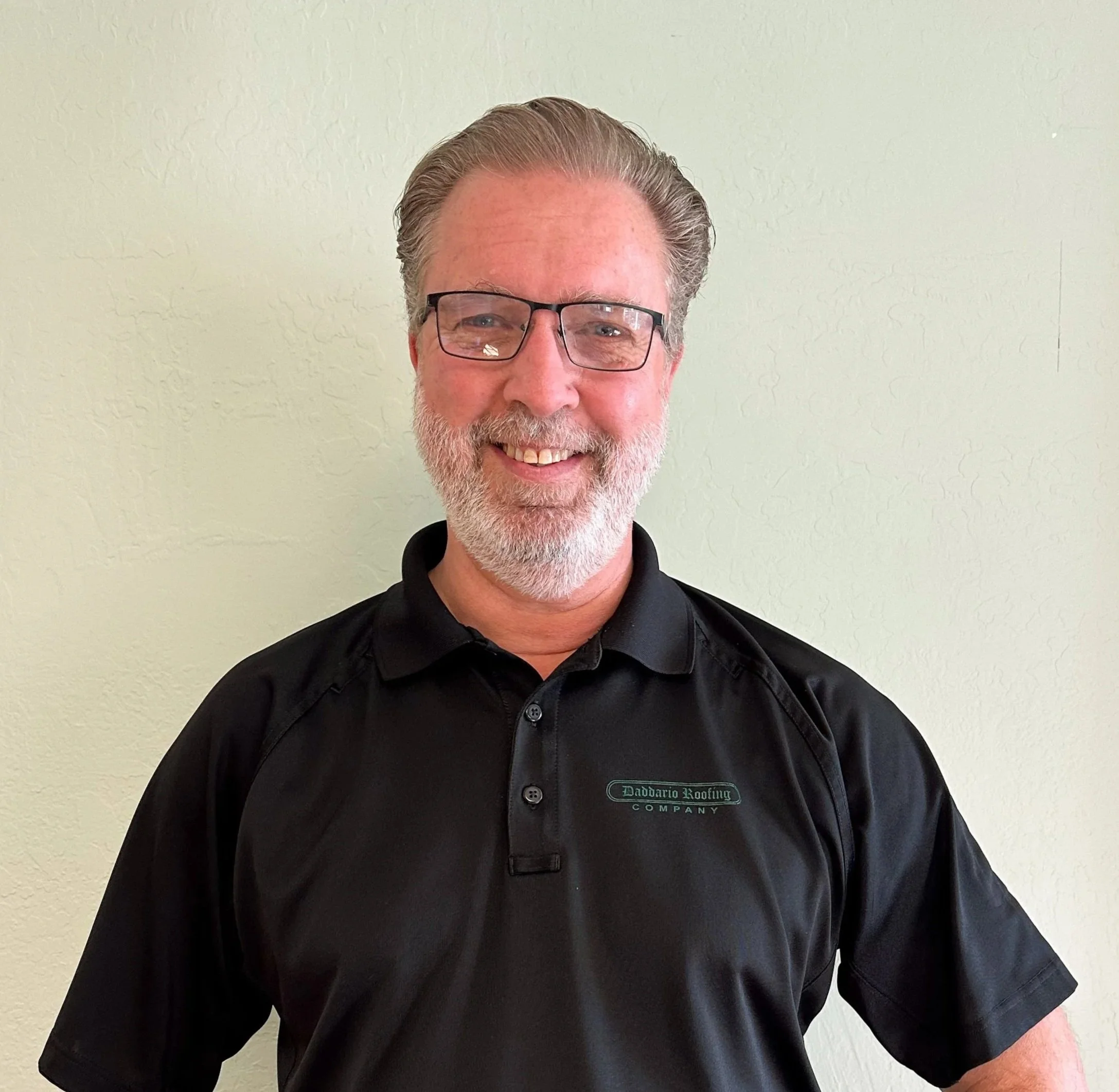 A smiling middle-aged man with gray hair and beard, wearing glasses, a black polo shirt with a company logo, blue jeans, and a smartwatch, standing against a light-colored wall.