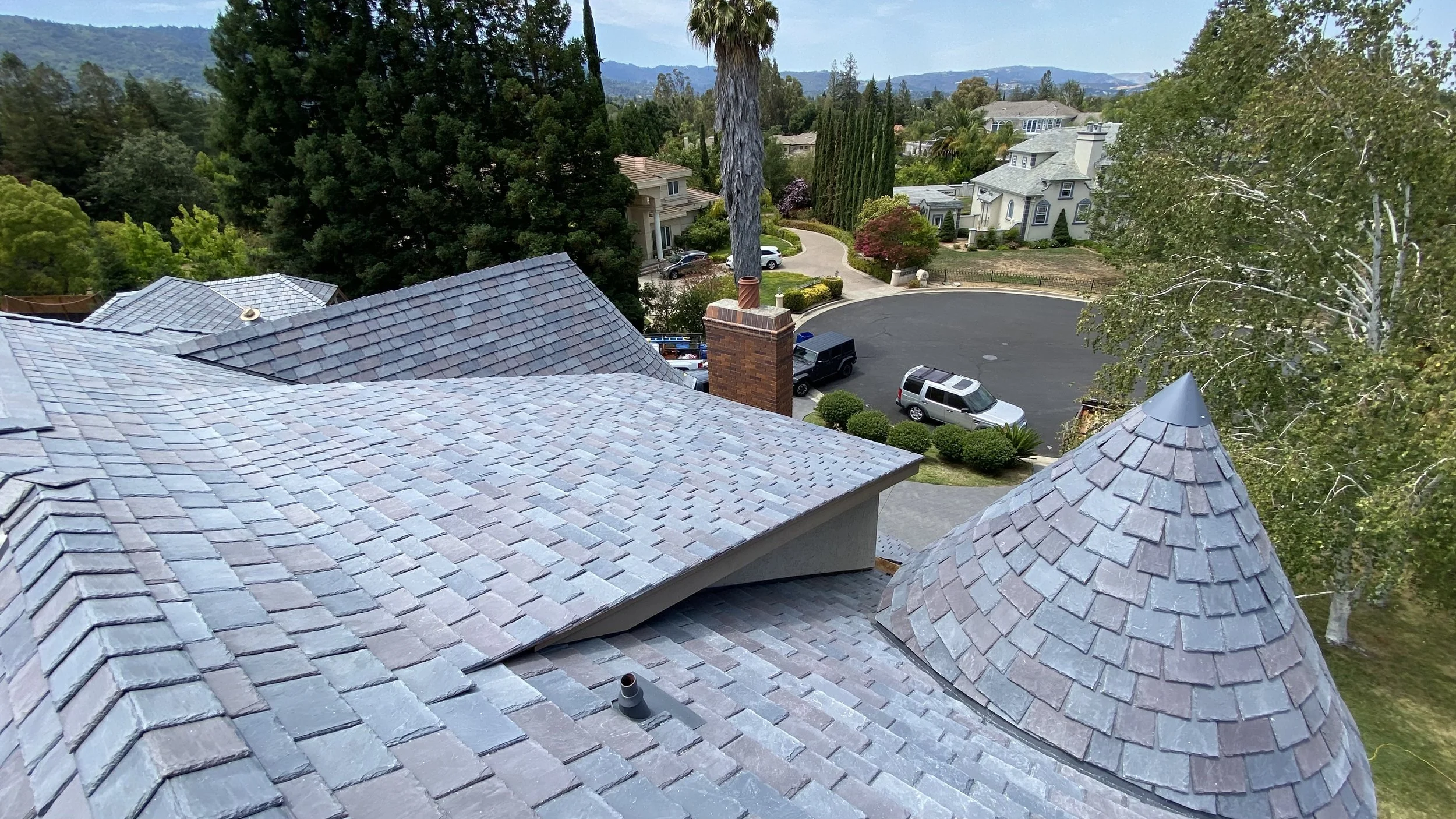 A residential neighborhood with houses, trees, and a street. The photo is taken from a high vantage point, showing rooftops made of gray and brown shingles, a chimney, and a conical turret on one house's roof.
