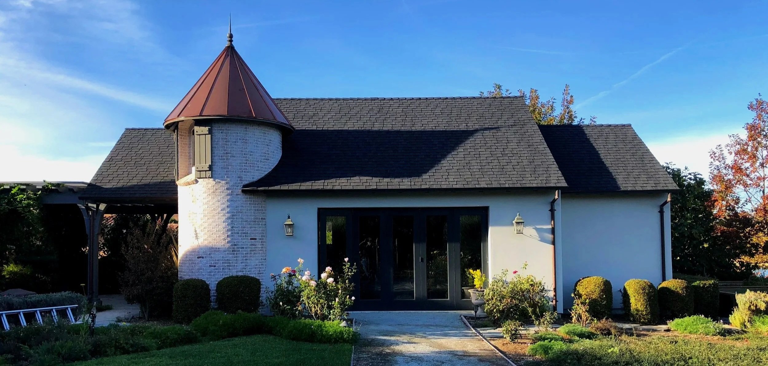 A house with a round turret and a pointed roof, white brick chimney, black doors, and surrounded by a manicured garden and lawn, under a blue sky.