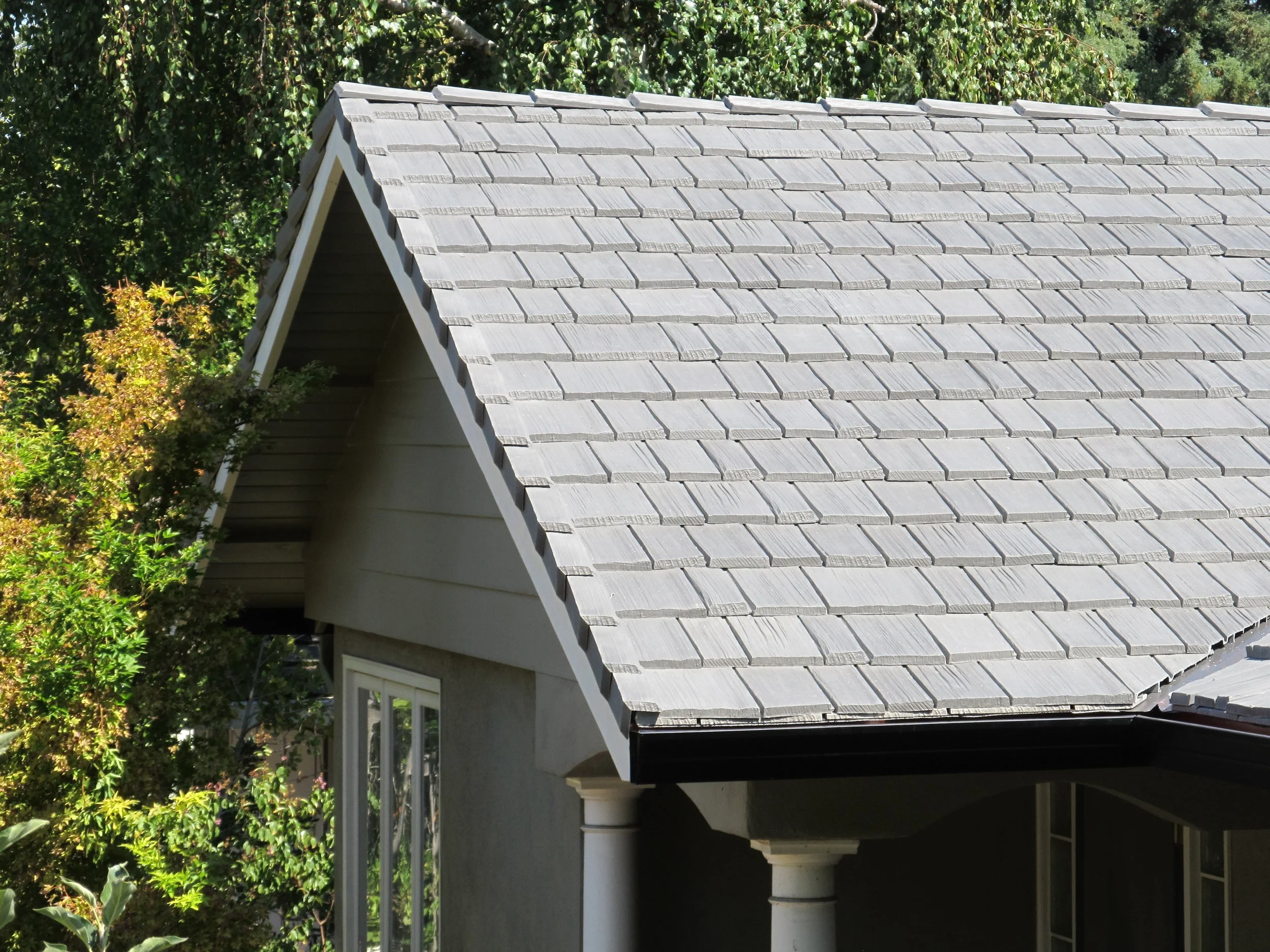Close-up view of a house roof with gray shingles, part of the house facade including a window and white columns, surrounded by green trees.