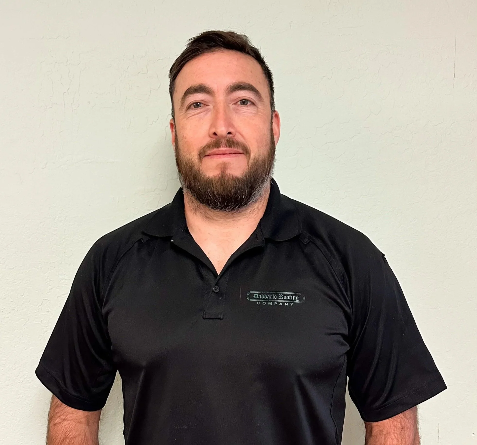 Man with brown hair and beard wearing a black polo shirt standing against a plain white wall.