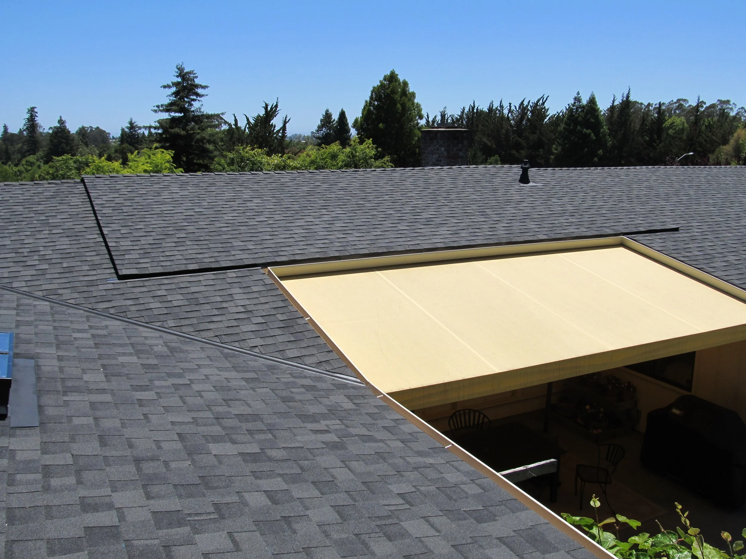A house roof with gray shingles, featuring a sunshade extension in a yard with trees in the background.