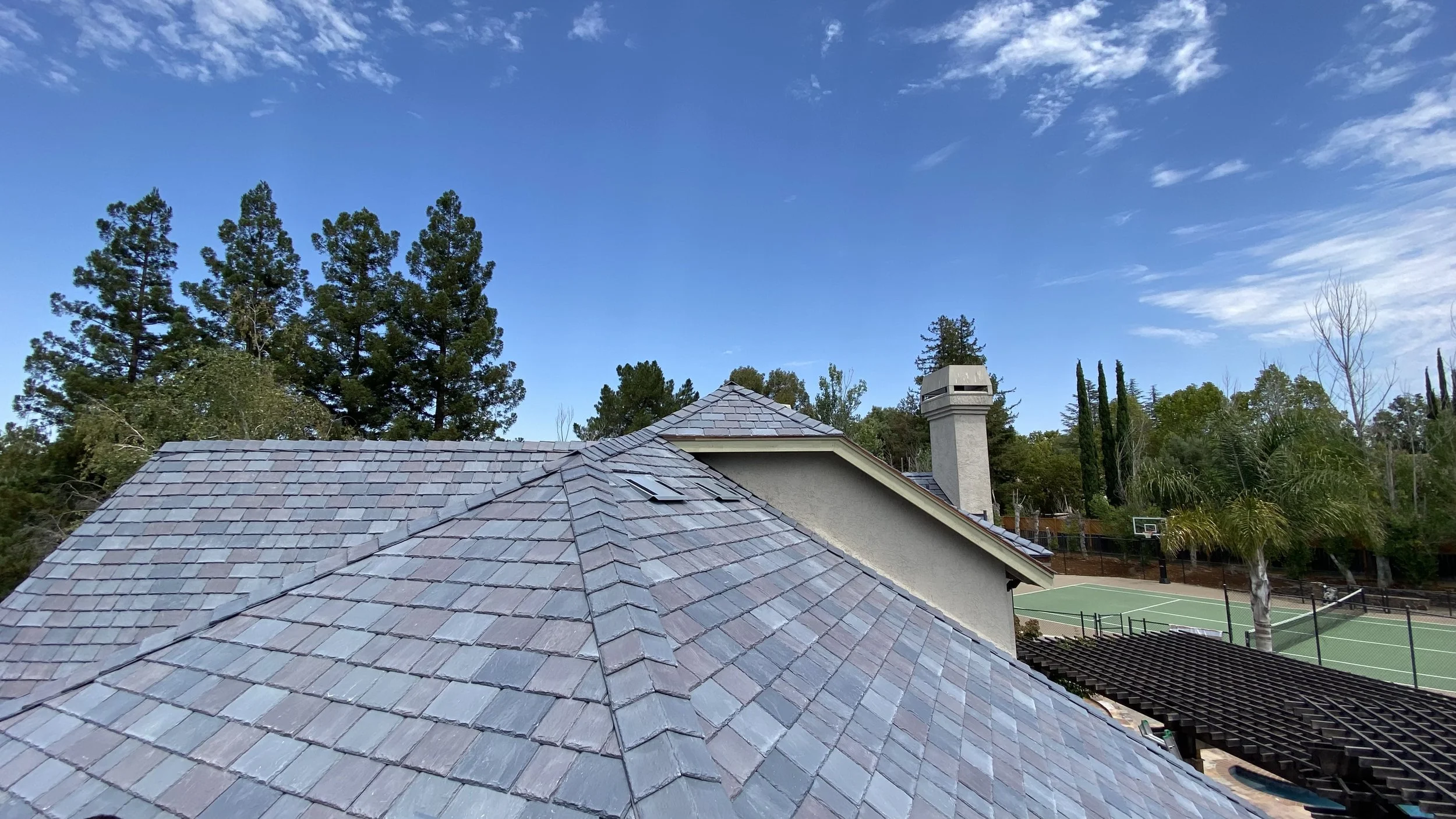 View of a residential house roof with gray shingles, a chimney, and a view of a backyard tennis court and trees under a blue sky with scattered clouds.