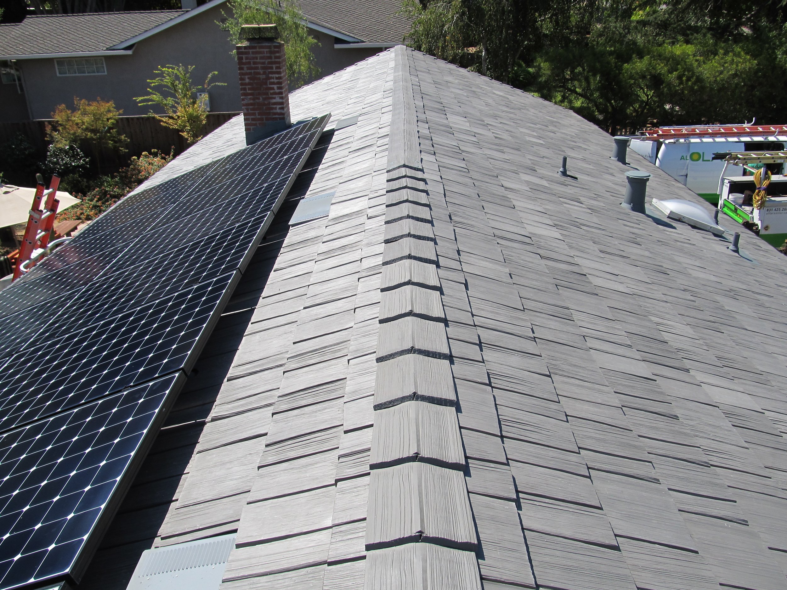 Rooftop with solar panels installed on it, visible chimney and multiple vents, surrounded by trees and neighboring houses.