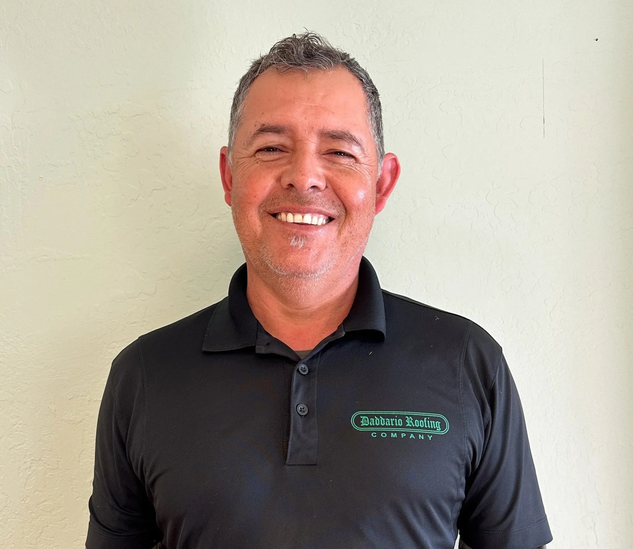 Smiling man wearing a black polo shirt with gray long sleeves underneath, standing against a light-colored wall.