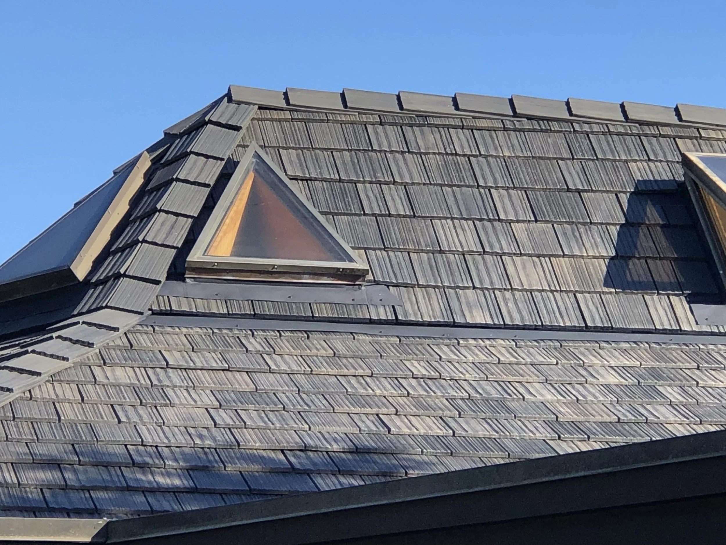 Close-up of a residential roof with wooden shingles and two triangular skylights, one with a wooden frame and one with a black frame, against a clear blue sky.