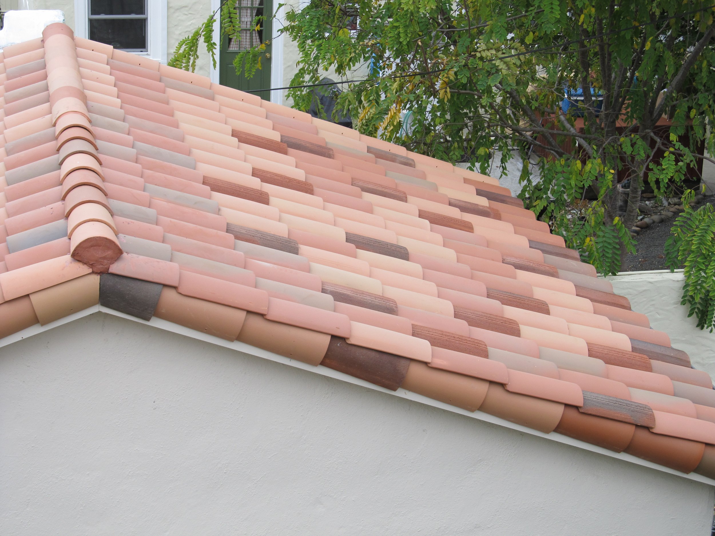 Close-up of a residential roof with pink and brown tiles, with greenery and a tree in the background.